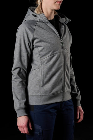 A person wearing an FXD Womens WF-3W Jacket, characterized by its light gray color and slightly textured zip-up design with a hood and pockets, is standing against a black background. The individual has blonde hair tied back and is dressed in dark pants. This casual, comfortable jacket offers both water resistance and windproof features.