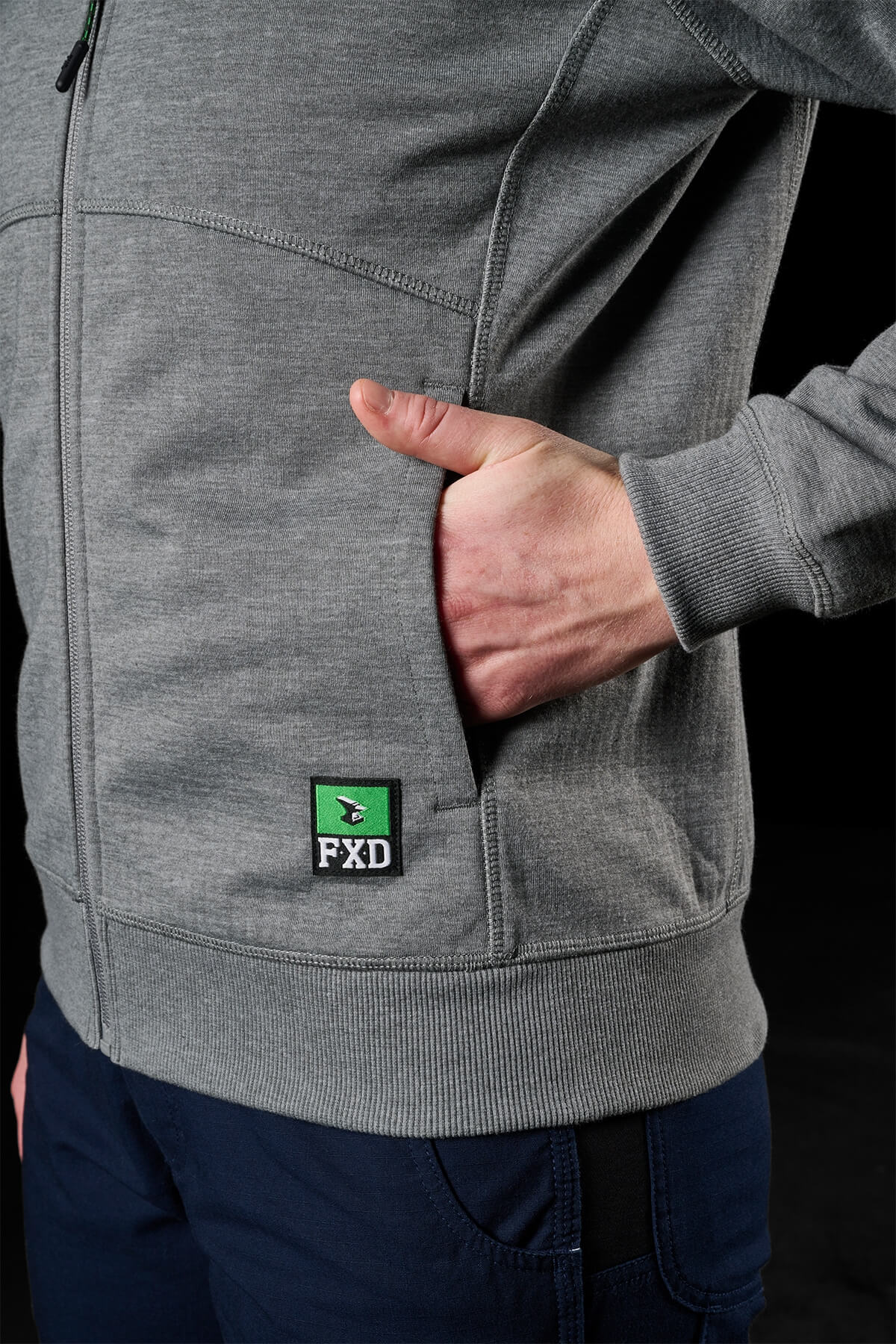 A person is dressed in an FXD Women's WF-3W Jacket, gray with a small green and black "FXD" logo near the pocket. Their right hand is casually placed in the jacket’s pocket, and they are wearing dark blue pants. The black background highlights the clothing's details, including its subtle windproof features.