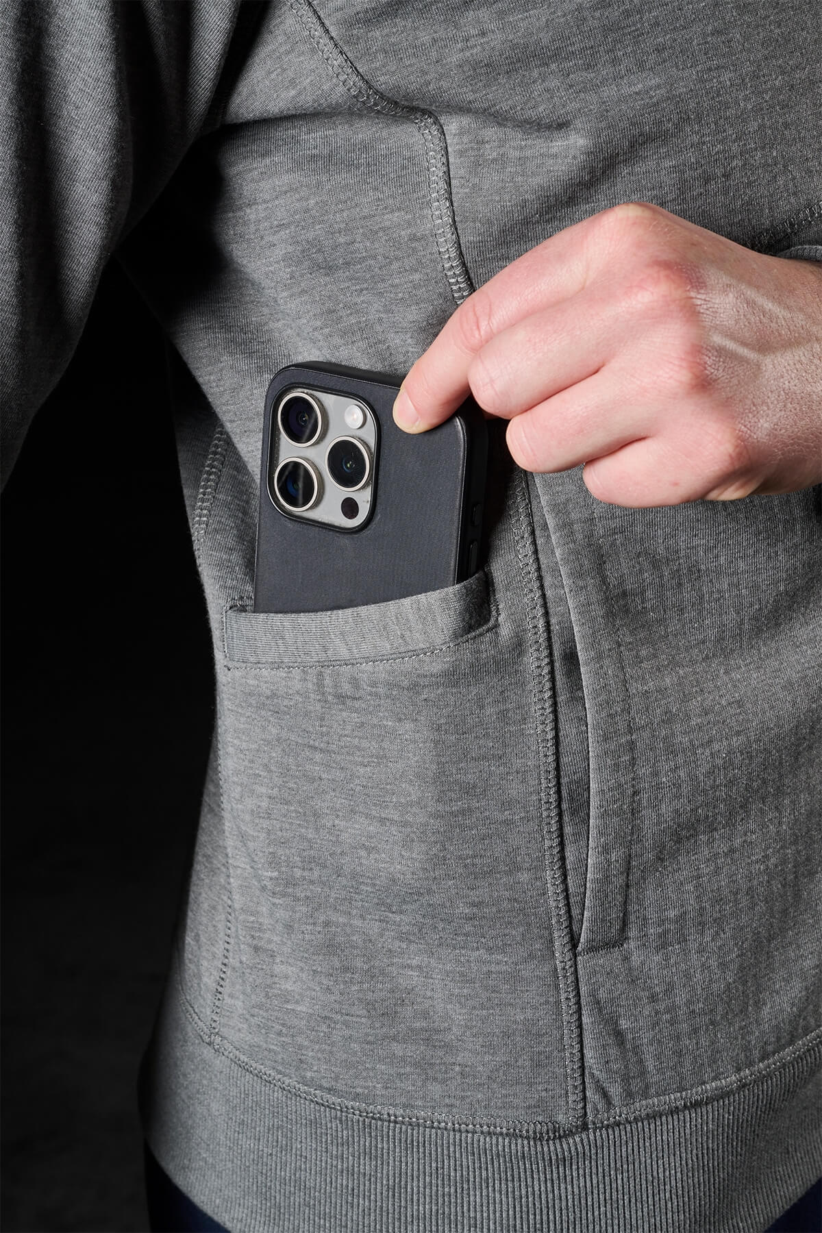 A person wearing an FXD Womens WF-3W Jacket in grey, which is windproof, places a smartphone with a black case into the jacket pocket. The phone's three camera lenses and flash are visible.