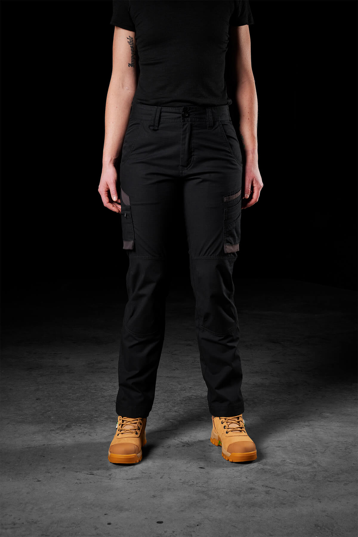 A person stands against a dark background, wearing black FXD Womens WP-7W cargo pants, a black shirt, and tan work boots. The person's arms are by their sides, and their face is not visible.