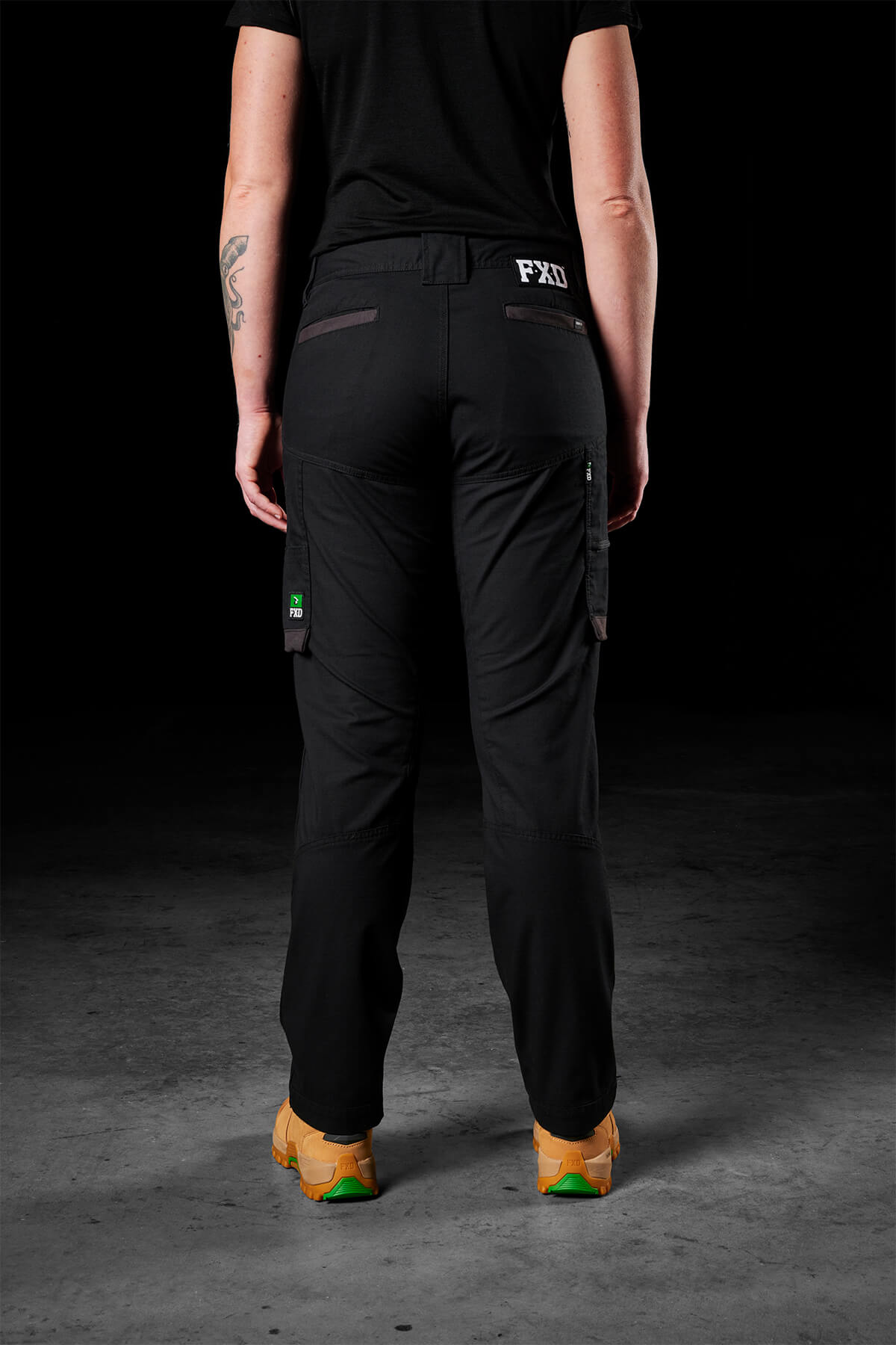 A person wearing black FXD Women's WP-7W cargo pants and tan shoes is shown from the back. The pants, crafted with flex double weave fabric and offering 50+ UPF protection, feature side and back pockets adorned with an "FXD" label. Dressed in a black shirt, they have a tattoo on their right forearm. The background is dark.