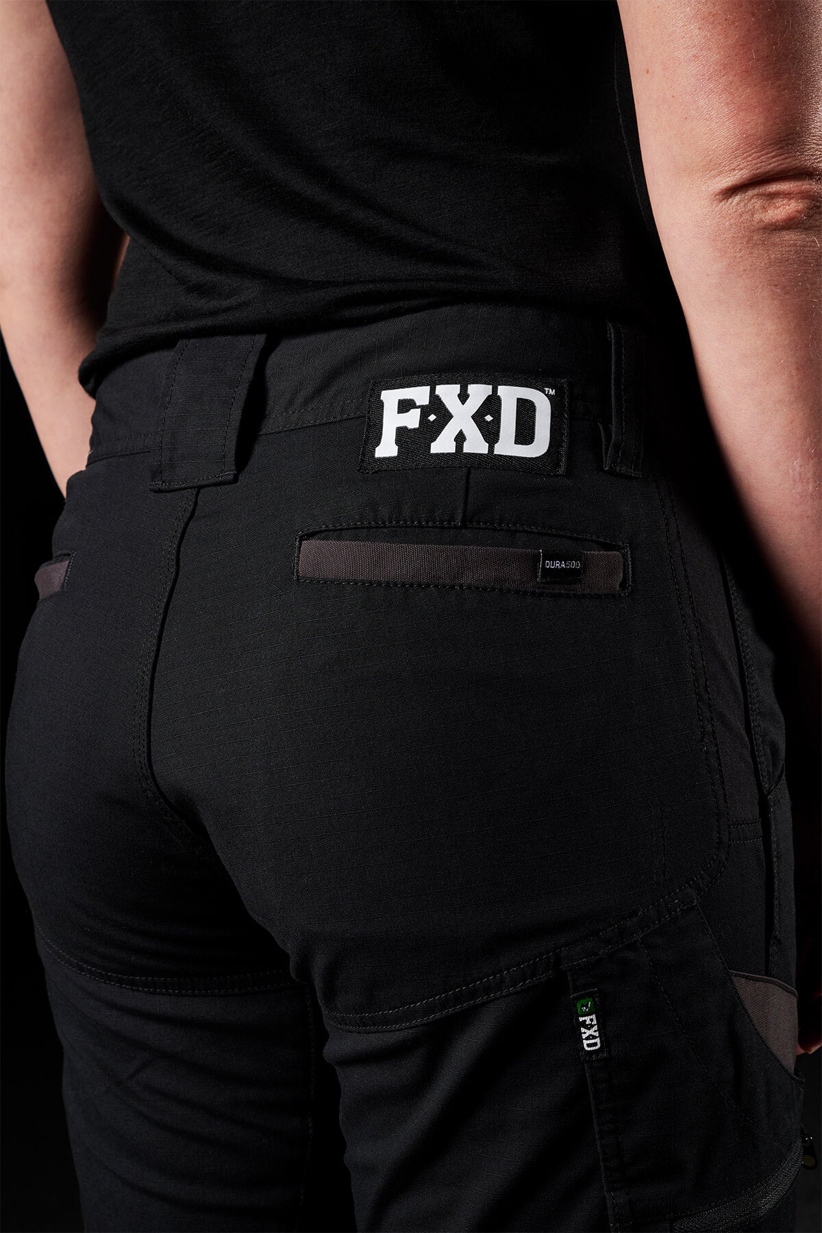 A person is wearing black FXD Womens WP-7W pants, embroidered with the "FXD" logo on the waist, shot from behind. The pants showcase pockets and detailed stitching and are crafted from flexible double weave fabric. They also don a black top, making their attire stand out against the dark background.