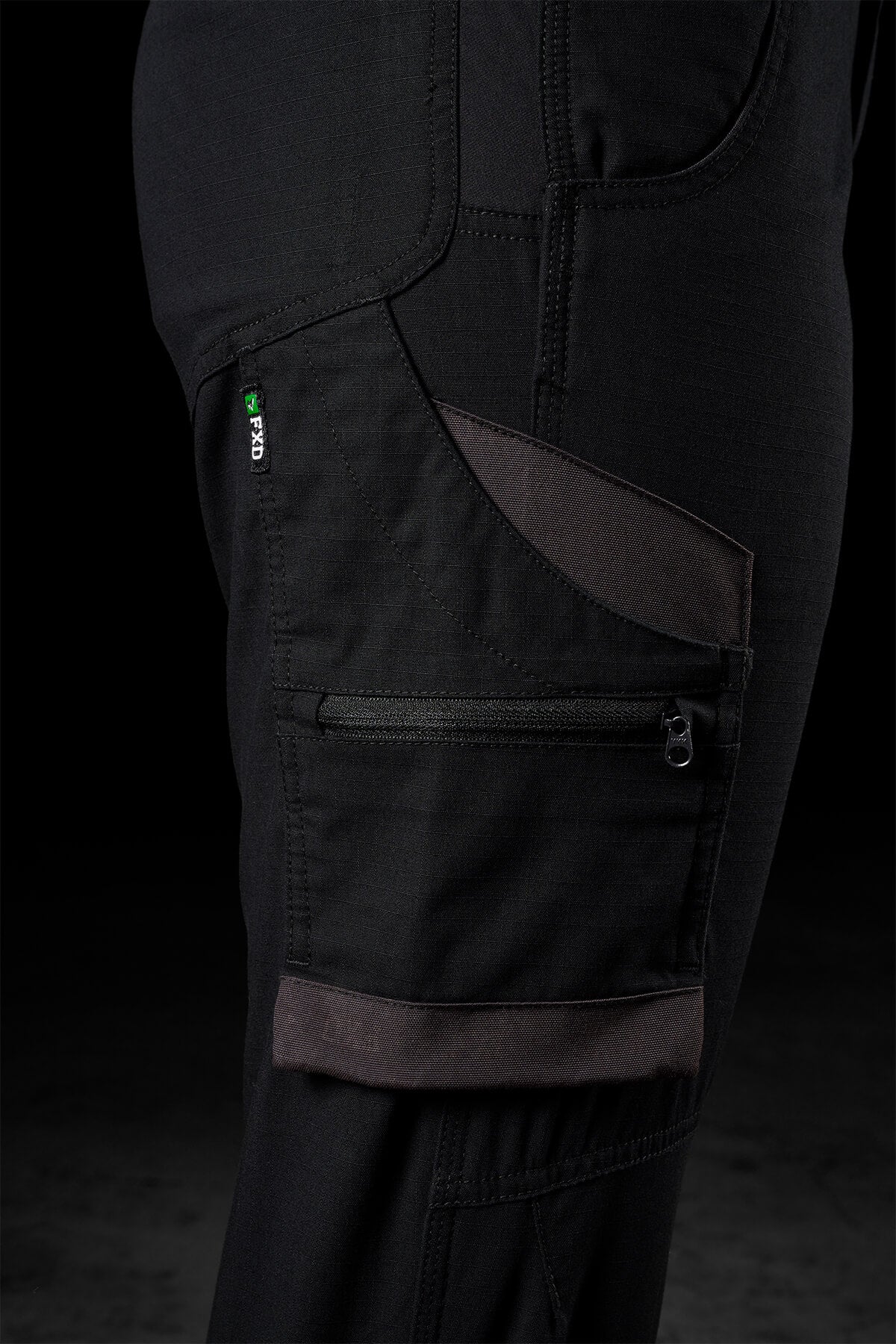 Close-up of a black, rugged FXD Womens WP-7W work pant with multiple pockets, including a zippered pocket and a smaller patch pocket. There is a green and white tag labeled "PRO" stitched near the top pocket. The stitching details, durable fabric, and flex double weave for 50+ UPF protection are visible.