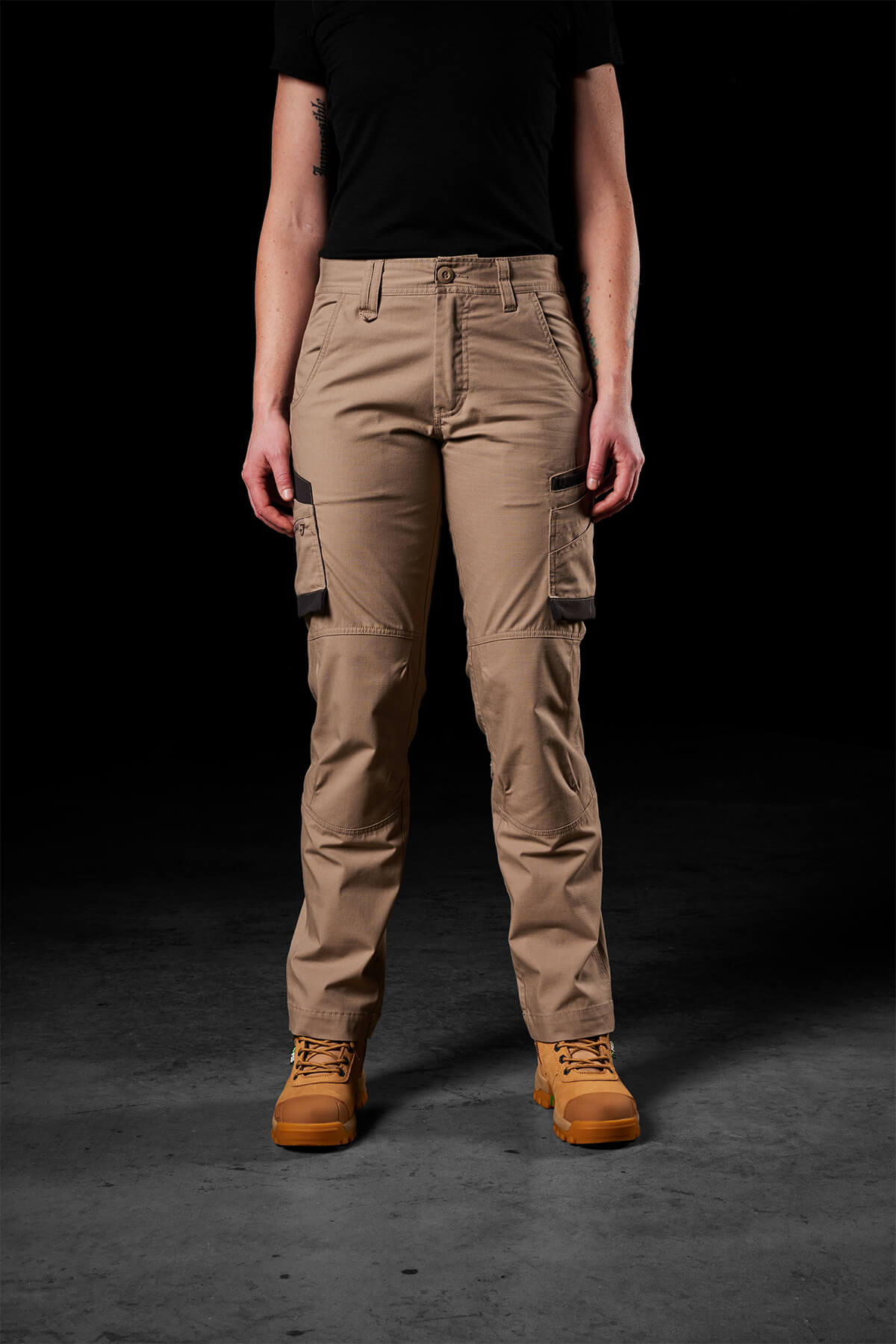 A person stands against a dark background, wearing FXD Womens WP-7W pants in black with multiple pockets and flex double weave hip panels, along with a black shirt and brown work boots. The focus is on the lower body from the mid-torso down. These pants offer 50+ UPF protection, making them ideal for any work environment.