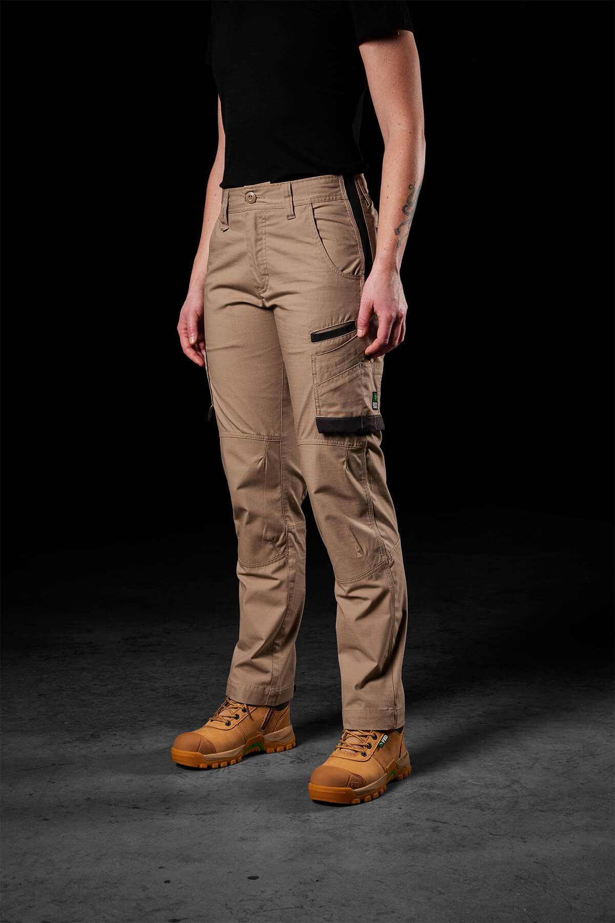 A person stands against a dark background, sporting a black short-sleeve shirt and FXD Womens WP-7W pants featuring khaki cargo styling and flex double weave hip panels. Their look is completed with tan work boots, though their face remains hidden in the image.