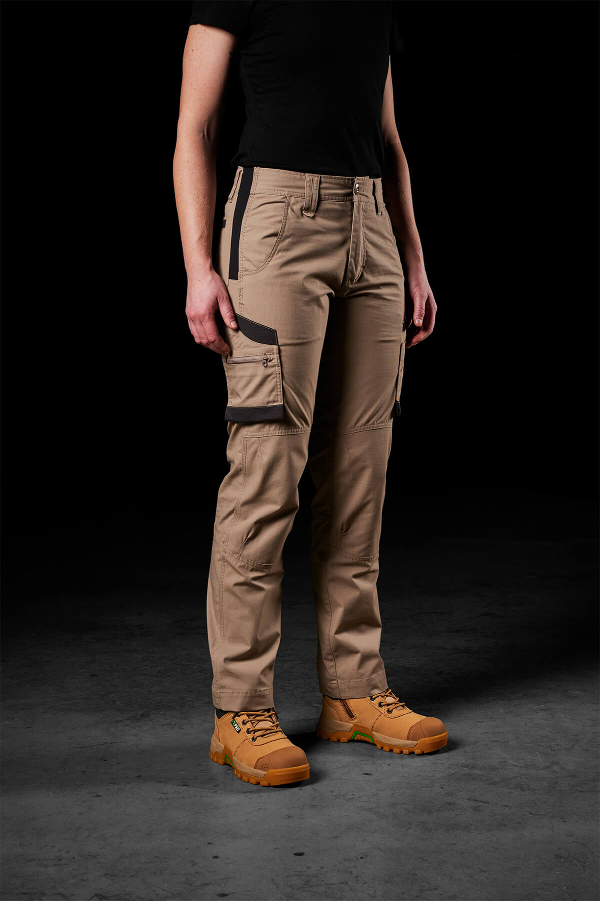 A person is standing against a dark background, wearing a black T-shirt, FXD Womens WP-7W pants featuring flex double weave hip panels and multiple pockets, and rugged tan boots. The person's head and hands are not visible in the frame.