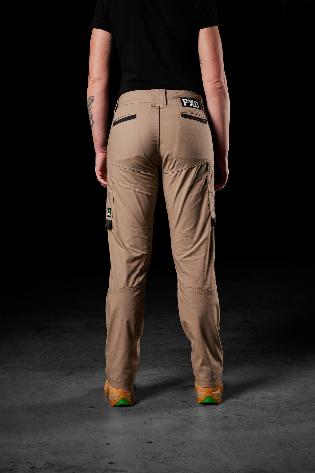 A person stands with their back to the camera, wearing a black shirt and FXD Womens WP-7W cargo pants in beige. These pants boast flex double weave hip panels and multiple pockets, including one adorned with a logo on the back. The dark background emphasizes the attire and its impressive 50+ UPF protection.