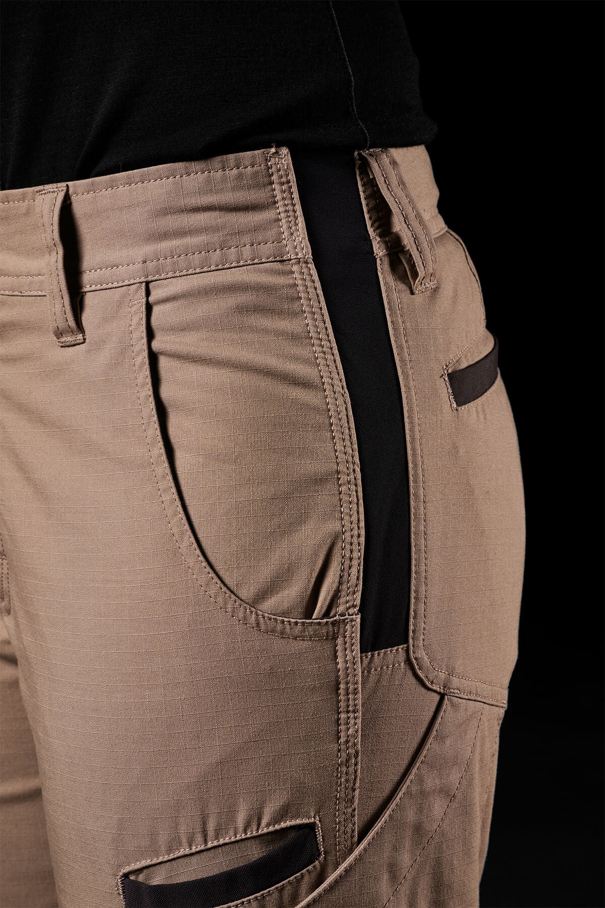Close-up of a person wearing FXD Womens WP-7W cargo pants in tan, showcasing black accents on the pockets and a black waistband. The durable fabric boasts flex double weave hip panels and offers 50+ UPF protection. These functional pants feature multiple pockets for convenience. The person is also dressed in a black top set against a dark background.