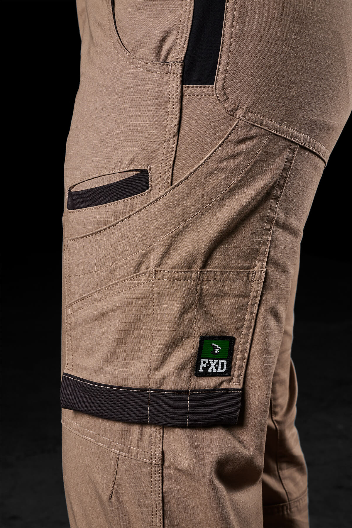A side view of a person wearing FXD Womens WP-7W tan pants showcases its multiple pockets and black accents. The design highlights include flex double weave hip panels, reinforced stitching, and a green FXD logo on one of the pockets. These pants also provide 50+ UPF protection. The backdrop is plain and dark.