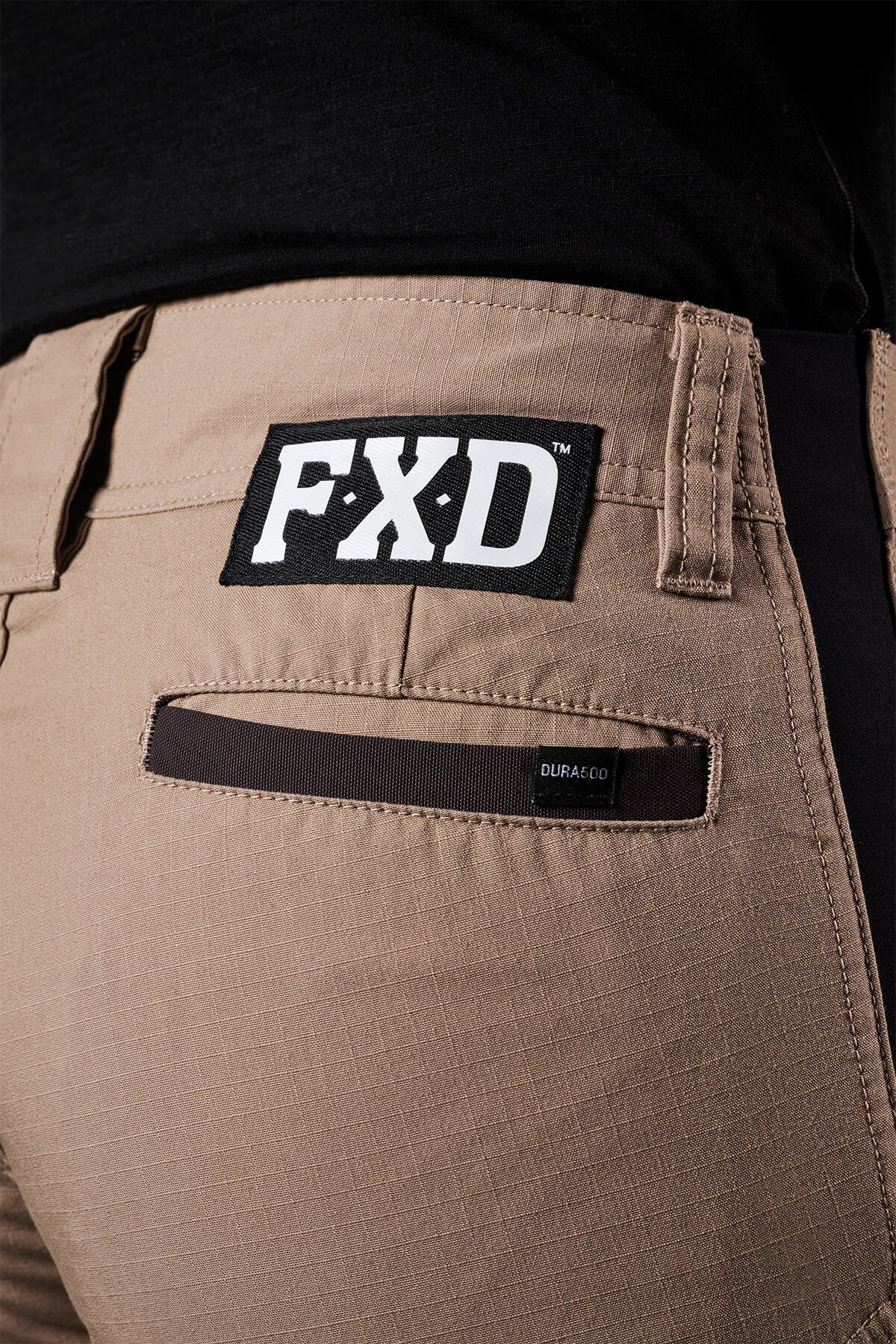 Close-up of FXD Womens WP-7W pants in beige, highlighting a black rectangular patch with white "FXD" lettering above the back pocket. The pocket also showcases a black strip labeled "DURA200." The wearer is dressed in a black top, accentuating the flex double weave hip panels designed for comfort.