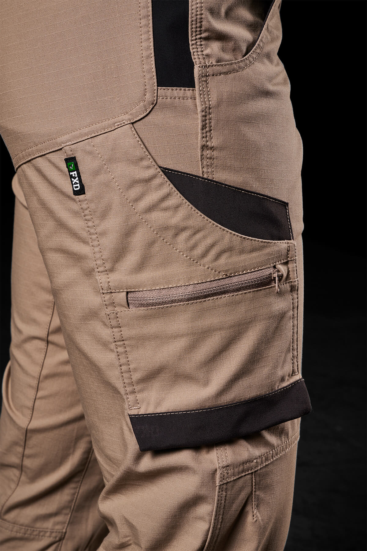 Close-up of a person wearing FXD Womens WP-7W pants in tan with black accents. These pants are designed with multiple pockets, including a zippered pocket and a compartment with a Velcro flap. They feature flex double weave hip panels for added durability and provide 50+ UPF protection. A small tag on the seam reads "FXD," and the dark background emphasizes the details of these stylish and functional pants.
