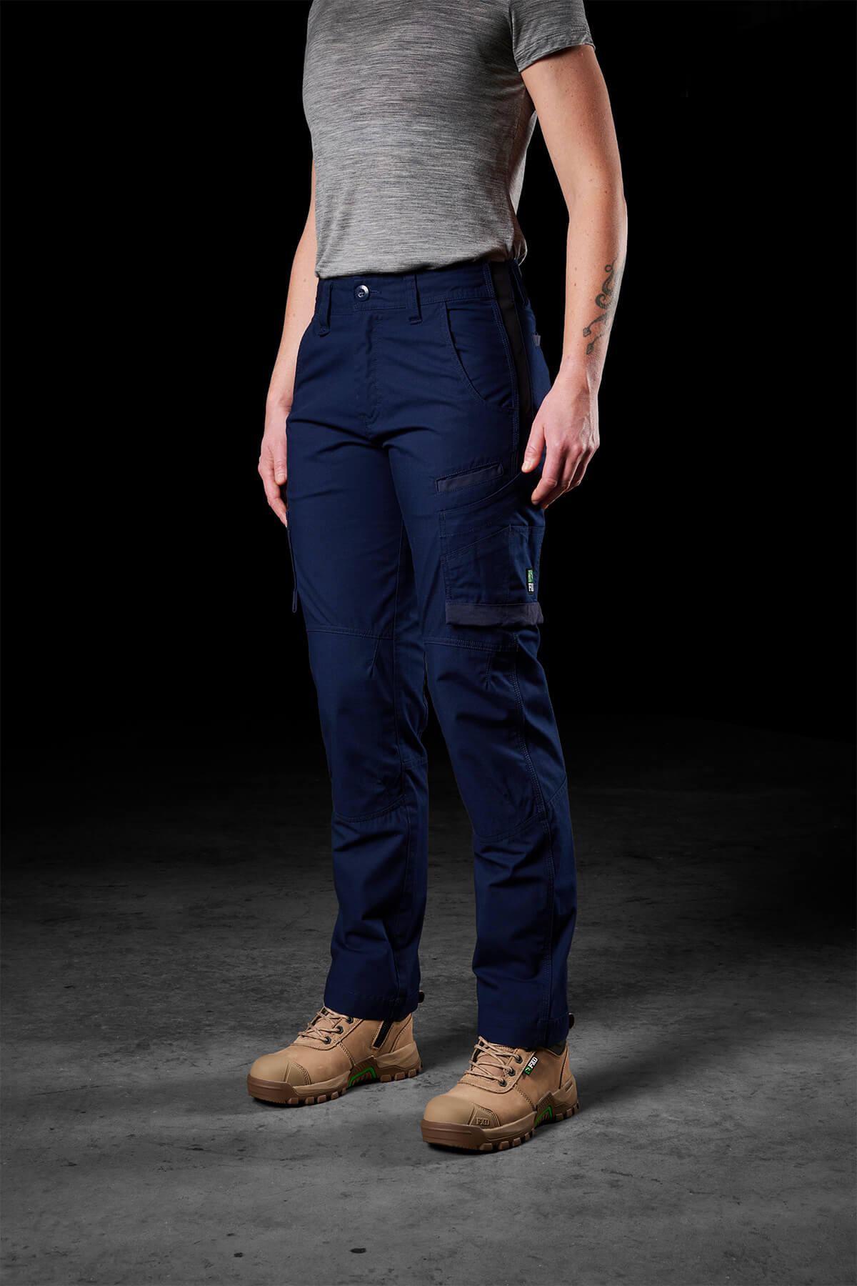 A person stands against a dark background, wearing a grey short-sleeved shirt, navy blue FXD Womens WP-7W pants featuring 50+ UPF protection, and beige work boots. Their hands are by their sides, revealing a tattoo on their left forearm.