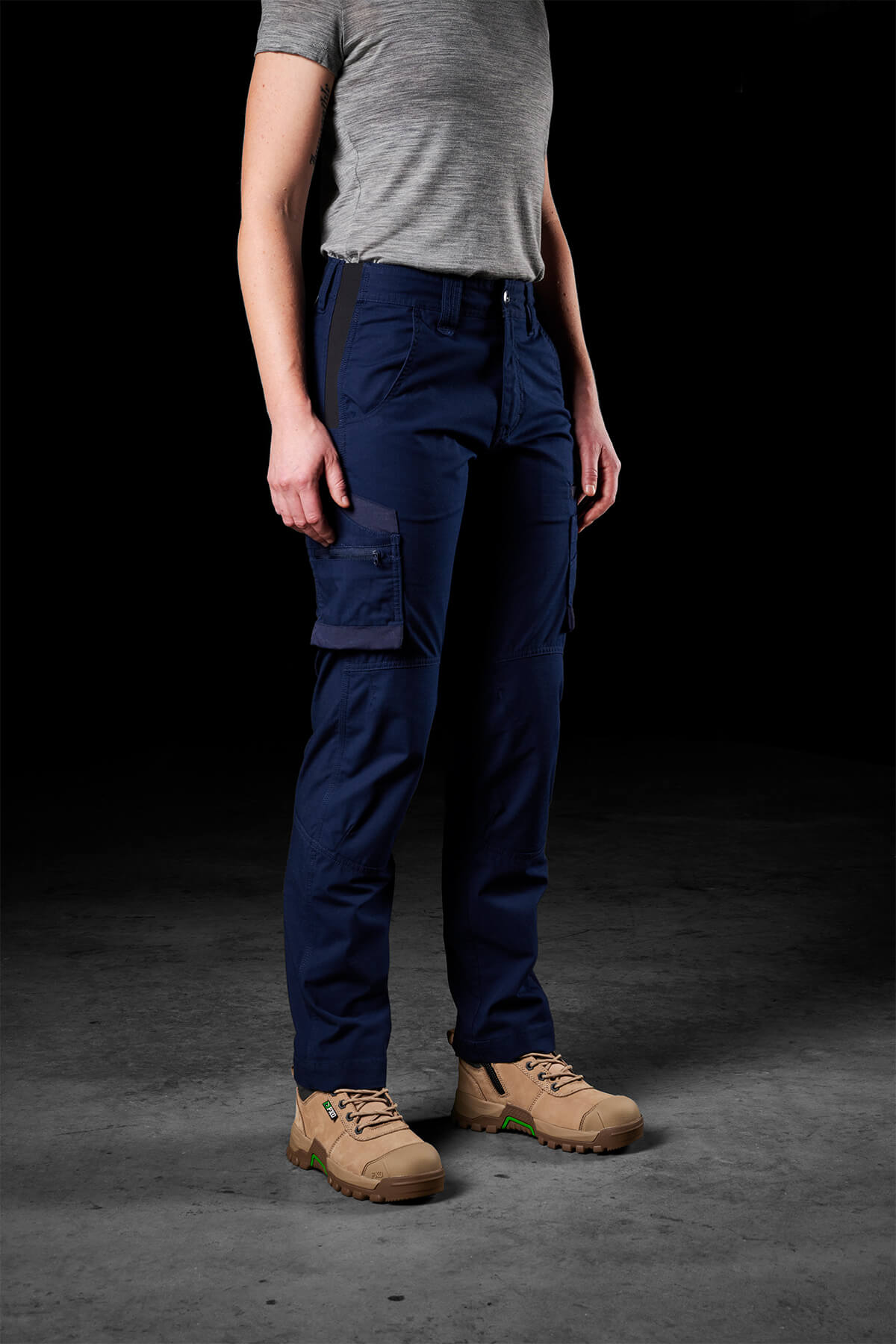 A person wearing a gray short-sleeve shirt, FXD Womens WP-7W navy blue cargo pants with flex double weave hip panels, and light tan hiking boots stands against a dark background. The person's face is not visible.