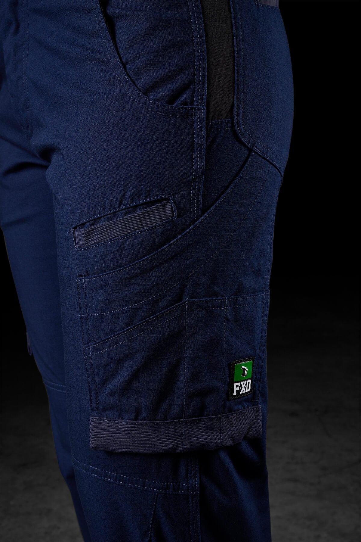 Close-up of FXD Womens WP-7W pants in dark blue, showcasing multiple pockets, a Velcro flap pocket, and a tool compartment. These work pants also feature reinforced stitching, flex double weave hip panels, and a small black and green FXD logo tag on the side. The background is dark and indistinct.