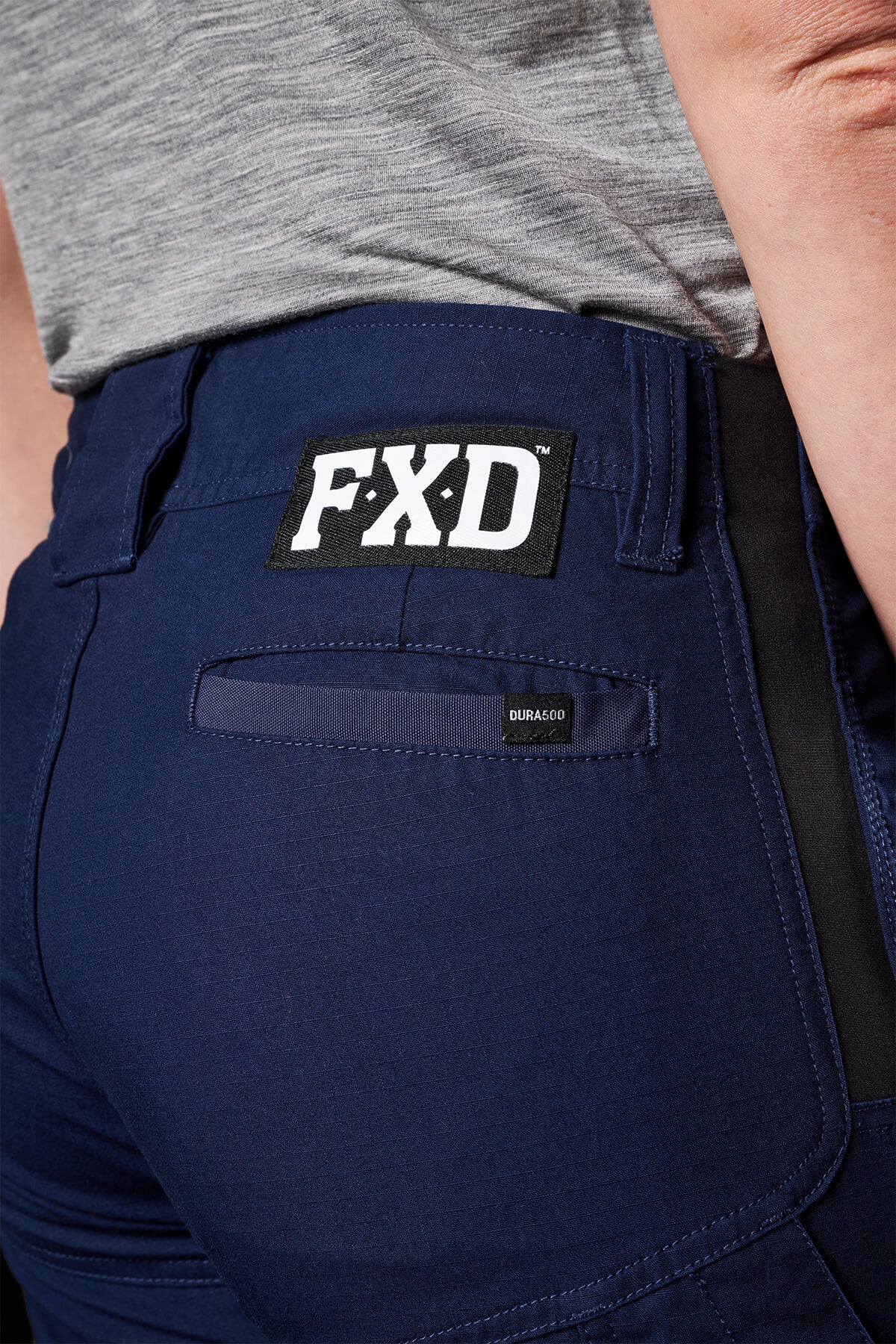 A close-up of a person wearing navy blue FXD Womens WP-7W pants with 50+ UPF protection and a black patch displaying "FXD" in white letters on the pocket area. The person's side and back are visible, along with their gray shirt. These pants also feature flex double weave hip panels and a small pocket with a "DURA500" tag.