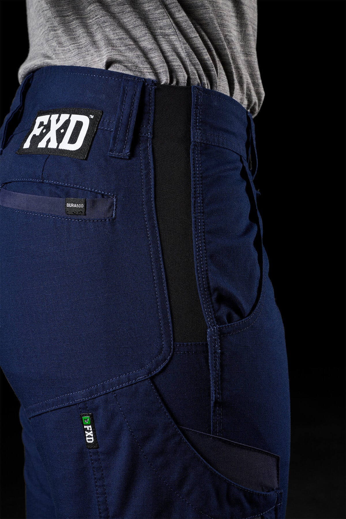 Close-up of a person wearing FXD Womens WP-7W pants in blue with an FXD logo on the back pocket, paired with a grey shirt tucked in. The image focuses on the waistband and side pockets, highlighting features such as 50+ UPF protection, flex double weave hip panels, precise stitching, high-quality material, and strategic logo placement against a dark background.