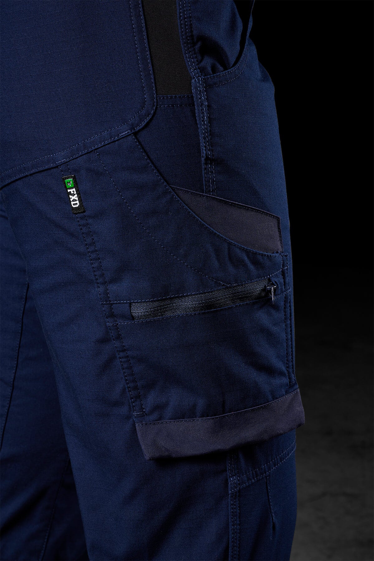 Close-up view of the side pocket on the FXD Womens WP-7W dark blue pants, featuring black accents on the pockets and knee area. The visibility tag labeled "PRO" and a zipper adorn one of the pockets. Flex double weave hip panels enhance durability, while the background remains dark and out of focus.