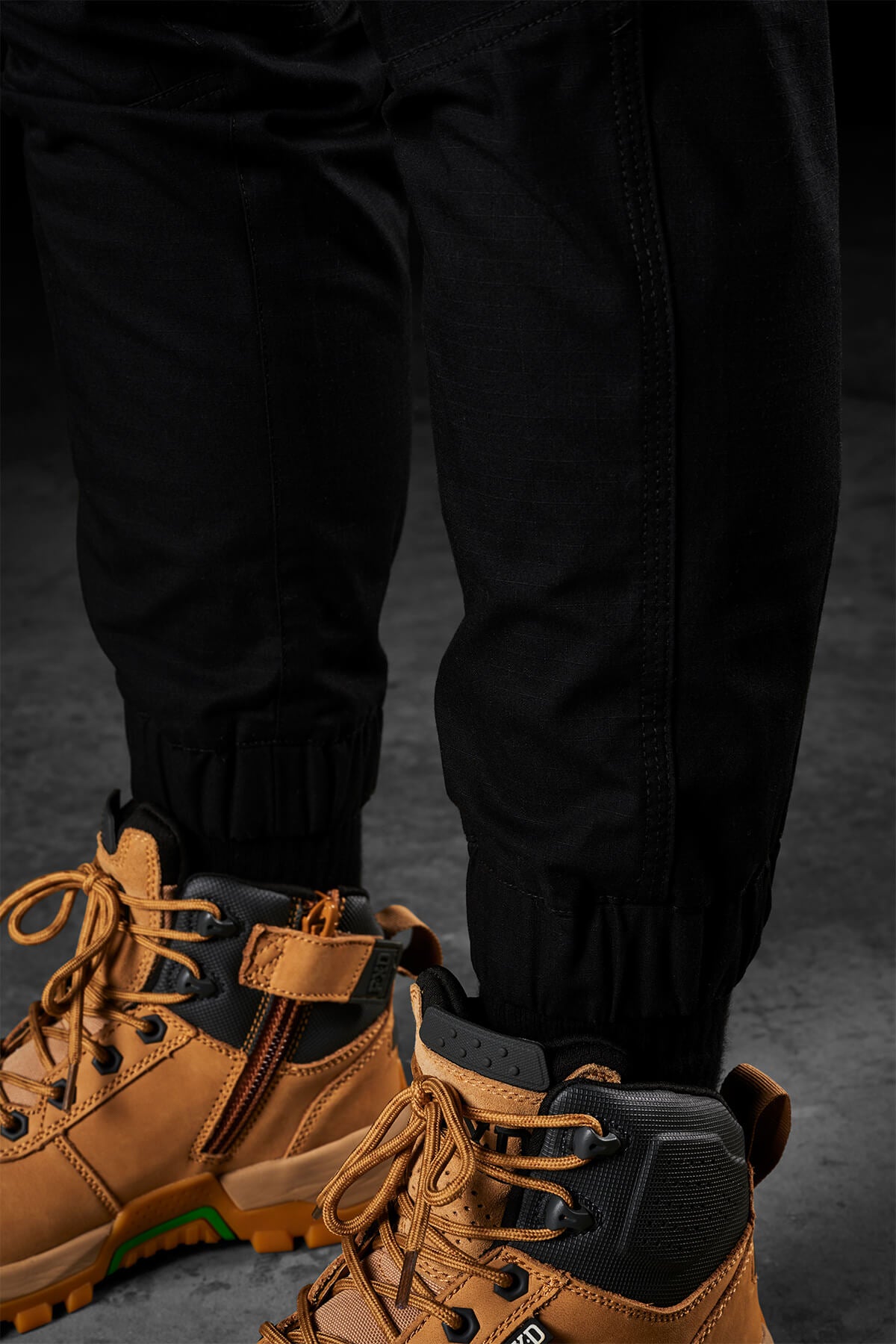 Close-up of a person wearing FXD Womens WP-8W black pants tucked into tan, high-top hiking boots with orange laces. The rugged boots feature zip and lace closures, while the lightweight stretch ripstop pant has elastic cuffs for mobility and comfort. The background is a dark, textured surface.