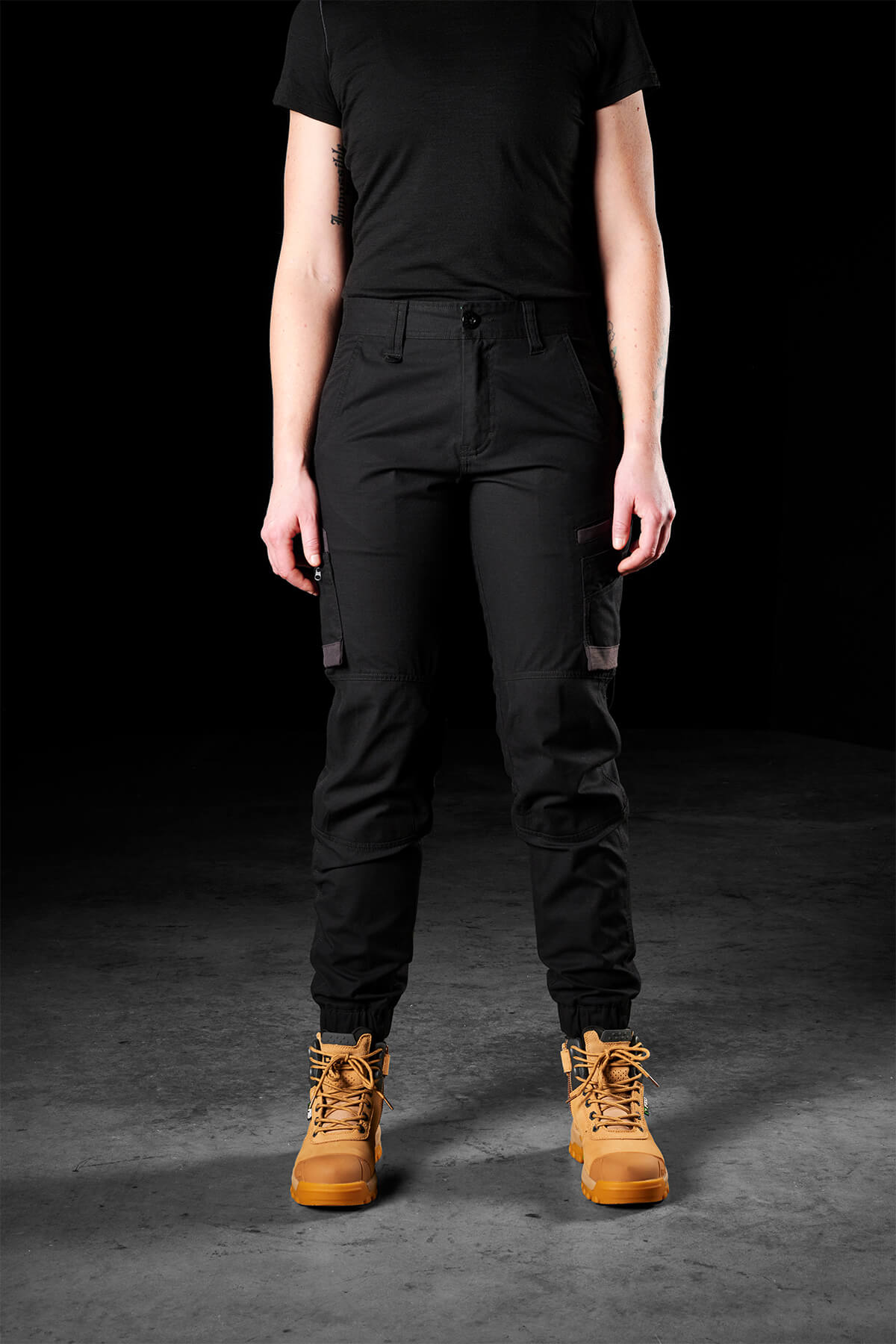A person stands against a dark background, wearing an FXD Womens WP-8W black short-sleeve shirt, lightweight stretch ripstop pants with abrasion panels, and tan work boots. Their arms are relaxed at their sides, and the focus is on their clothing and boots.