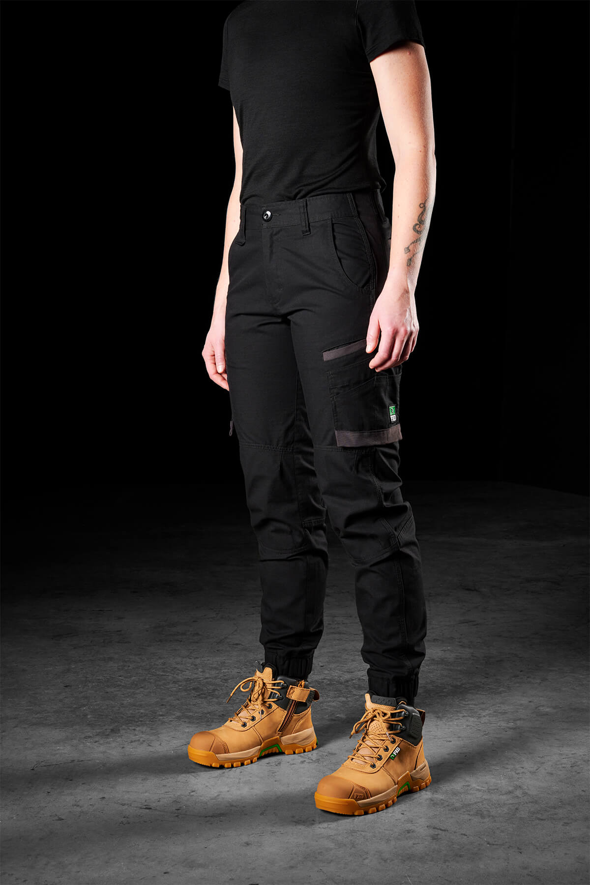 A woman dressed in the FXD Womens WP-8W shirt and lightweight stretch ripstop pants stands on a dark, textured floor. The outfit is paired with tan work boots that have dark laces and rugged soles. Her right arm is visible, showing a tattoo. The background is black.