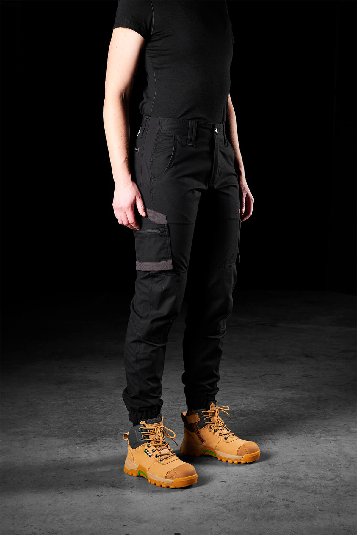 A person standing against a dark background is wearing black FXD Womens WP-8W tactical pants with multiple pockets and tan lace-up boots. The lightweight stretch ripstop FXD Womens WP-8W pants feature abrasion panels for durability. The person is also wearing a black, short-sleeved shirt, with their upper body not visible in the frame. The floor appears to be a solid, dark surface.