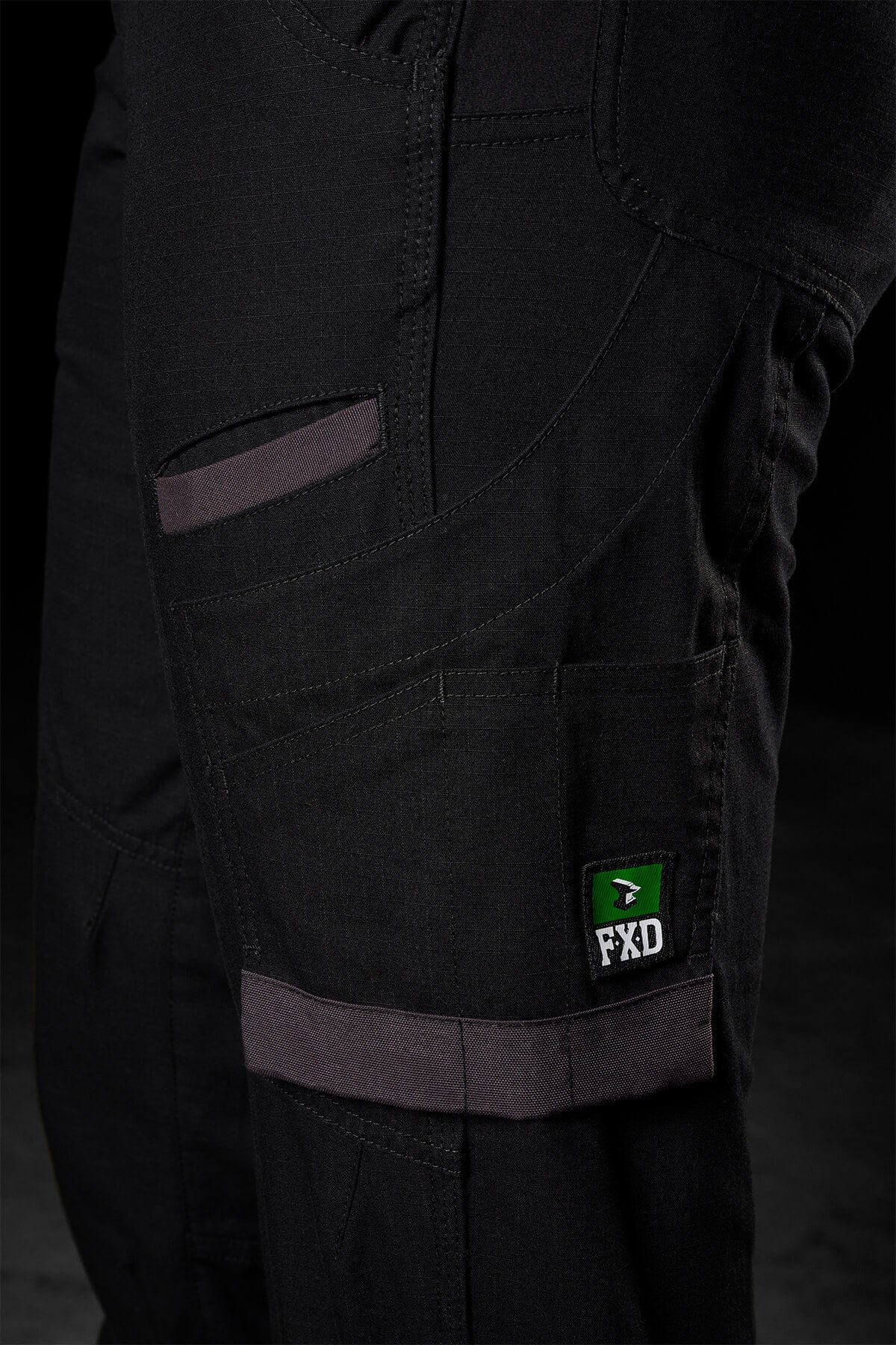 Close-up of a pair of FXD Womens WP-8W black pants showcasing multiple pockets, reinforced stitching, and a distinctive green label with the text "FXD." The gray-accented pockets and durable appearance indicate practicality. These lightweight stretch ripstop pants also feature abrasion panels, making them ideal for work or outdoor activities.