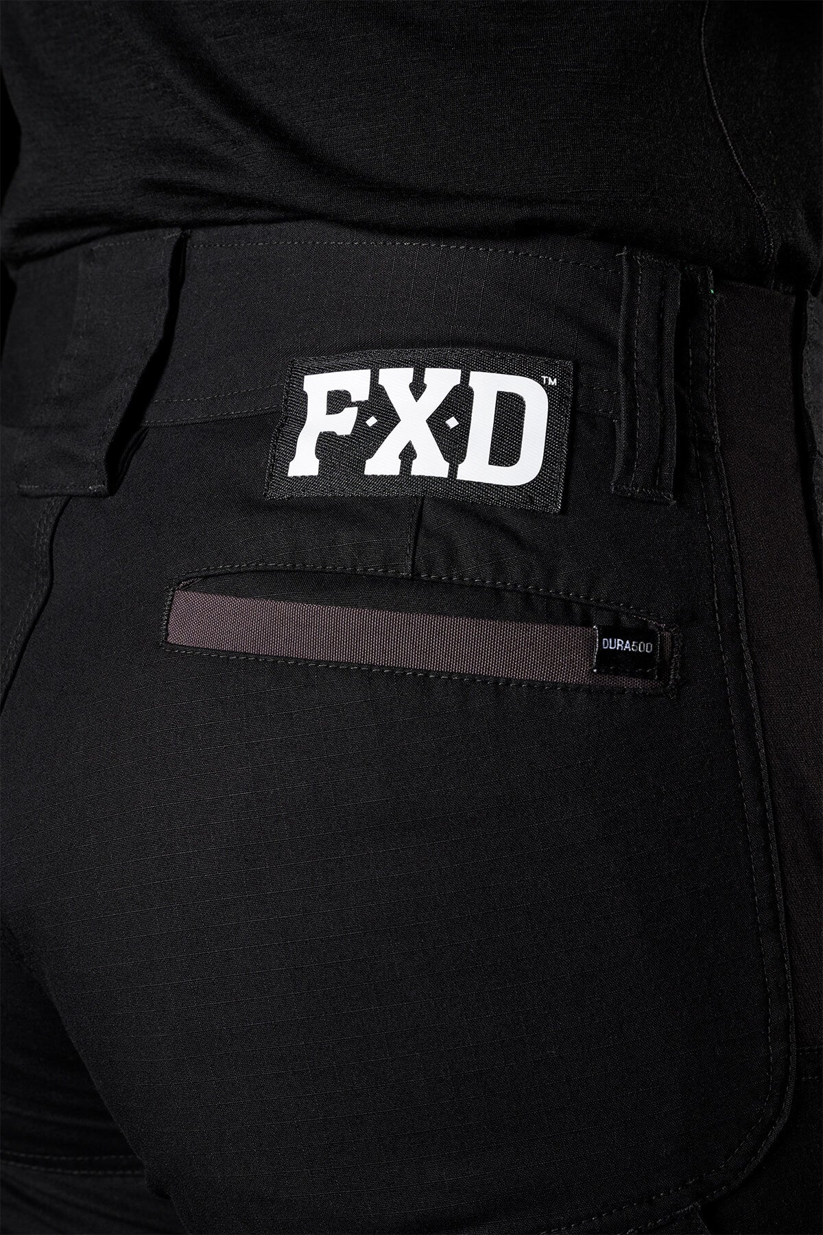 Close-up of a person wearing FXD Womens WP-8W black lightweight stretch ripstop pants, with a visible FXD logo on the back pocket. The pants also feature a small label next to the pocket and 50+ UPF protection. The image focuses on the back part, showcasing the fabric, stitching details, and abrasion panels.