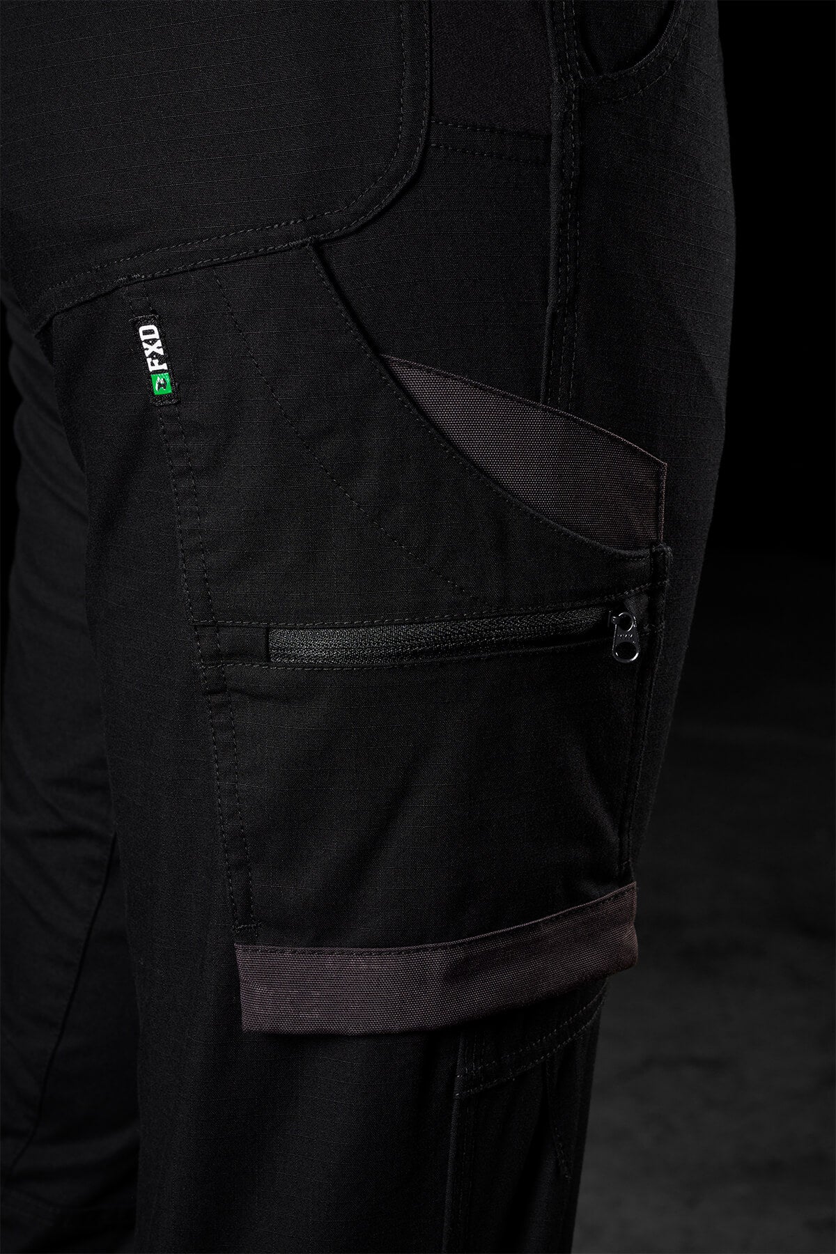 Close-up of a black zippered utility pocket on the FXD Womens WP-8W lightweight stretch ripstop pants, showcasing the detailed stitching and fabric texture. The pants feature additional small pockets and a small green and white tag with text near the pocket seam. The background is dark and out of focus.
