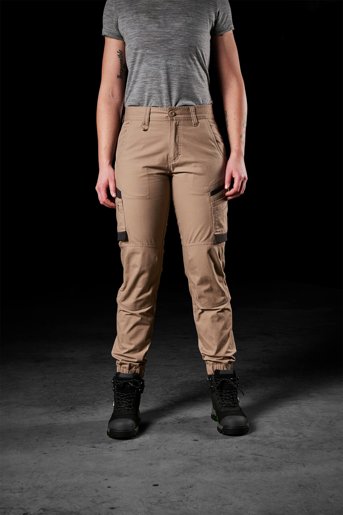 A person stands facing forward wearing a gray t-shirt, FXD Womens WP-8W lightweight stretch ripstop pants with multiple pockets for maximum range of movement, and black boots. The background is dark, making the clothing stand out.