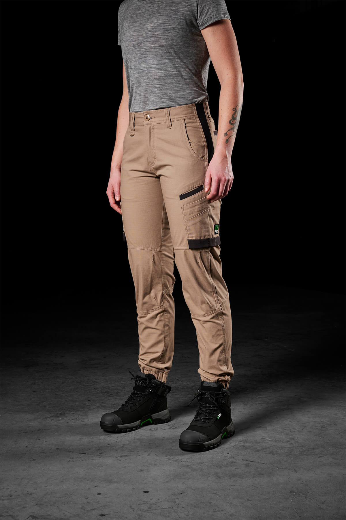A person wearing a gray short-sleeve shirt, FXD Womens WP-8W tan lightweight stretch ripstop pants with zippered pockets and 50+ UPF protection, and black work boots stands against a dark background. The person's right arm is visible, showing a tattoo.