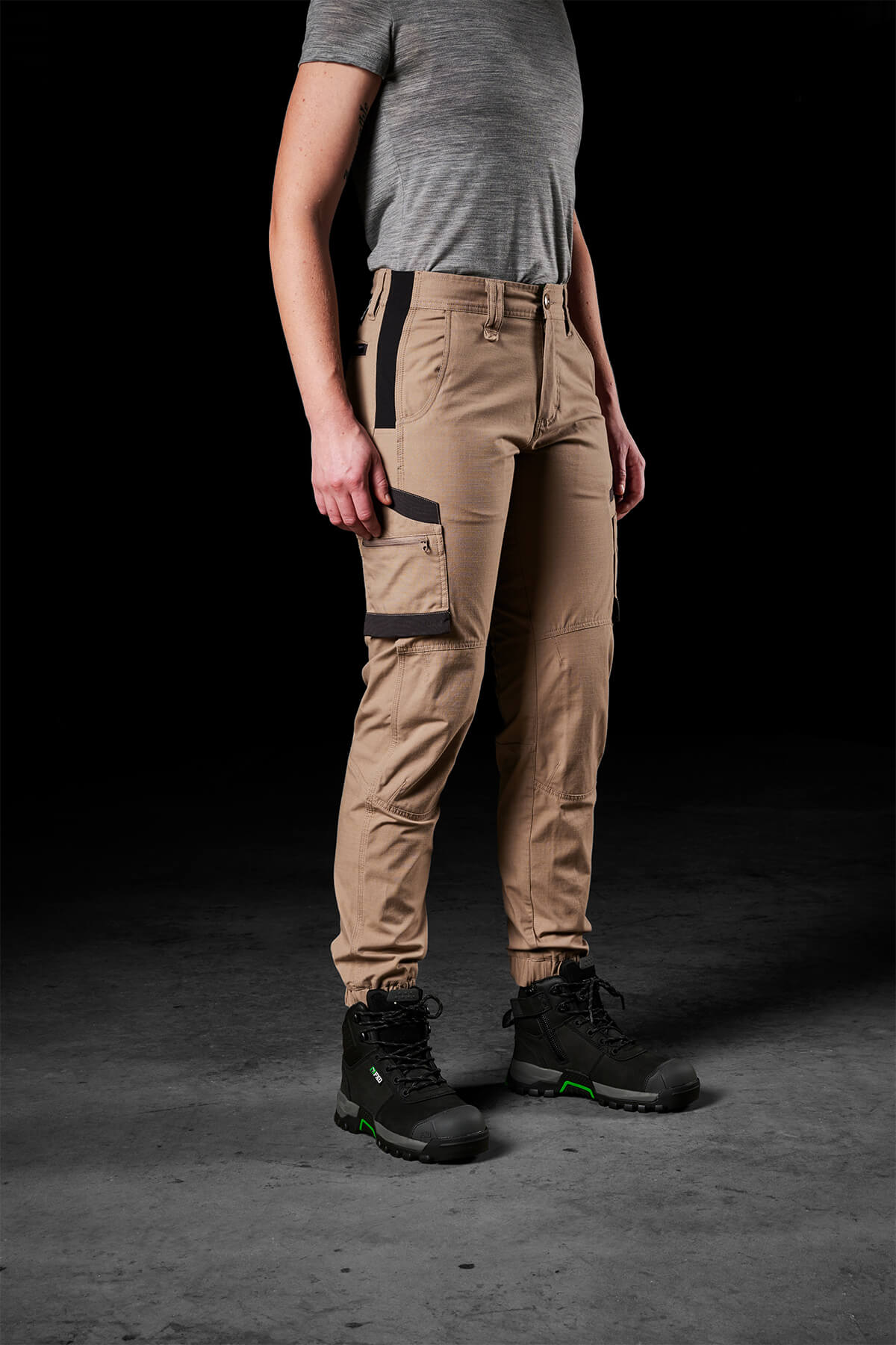 A person wearing a gray t-shirt and FXD Womens WP-8W beige cargo pants with black pockets, paired with black and green hiking boots, stands against a dark background. These lightweight stretch ripstop pants have multiple pockets and offer maximum range of movement, making the attire practical for outdoor activities.