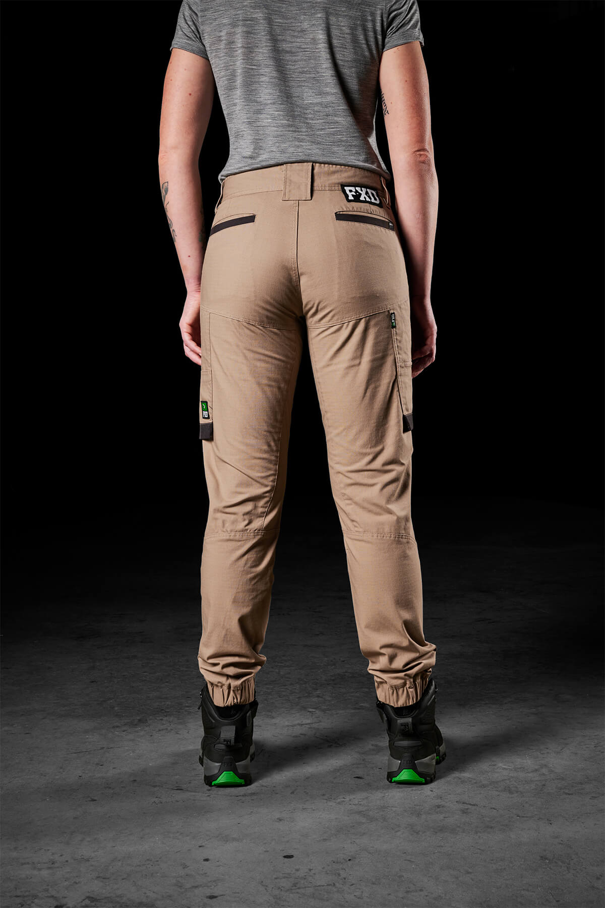 A person is standing with their back facing the camera, wearing a gray shirt and FXD Womens WP-8W beige lightweight stretch ripstop pants featuring pockets and black accents. They are also wearing dark-colored shoes with green soles. The dark and neutral background enhances the focus on their attire, which offers maximum range of movement.