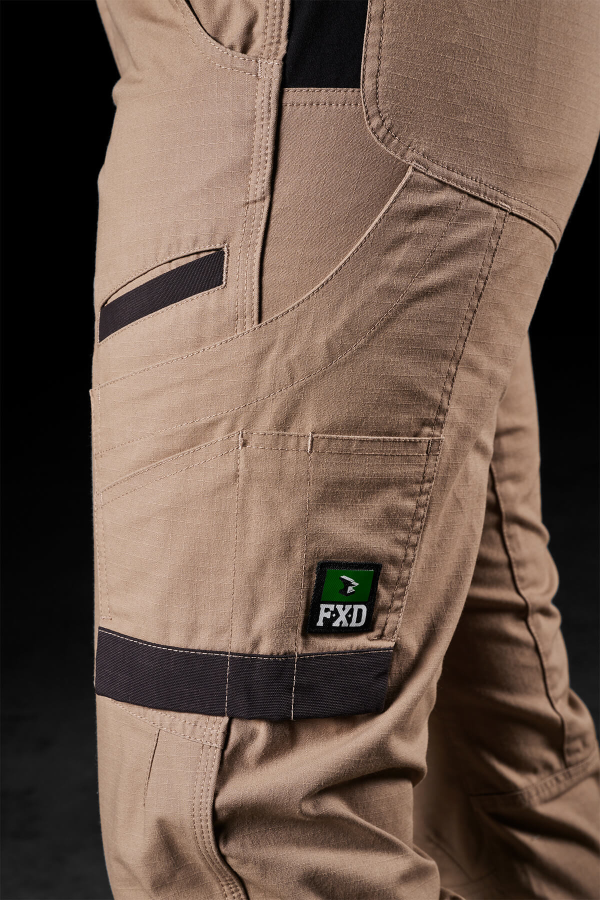 Close-up of a person wearing FXD Womens WP-8W beige work pants with black accent patches. These lightweight stretch ripstop pants feature multiple pockets and a green and black "FXD" brand logo on the side. The background is black, emphasizing the detail and texture of the fabric, designed for maximum range of movement.