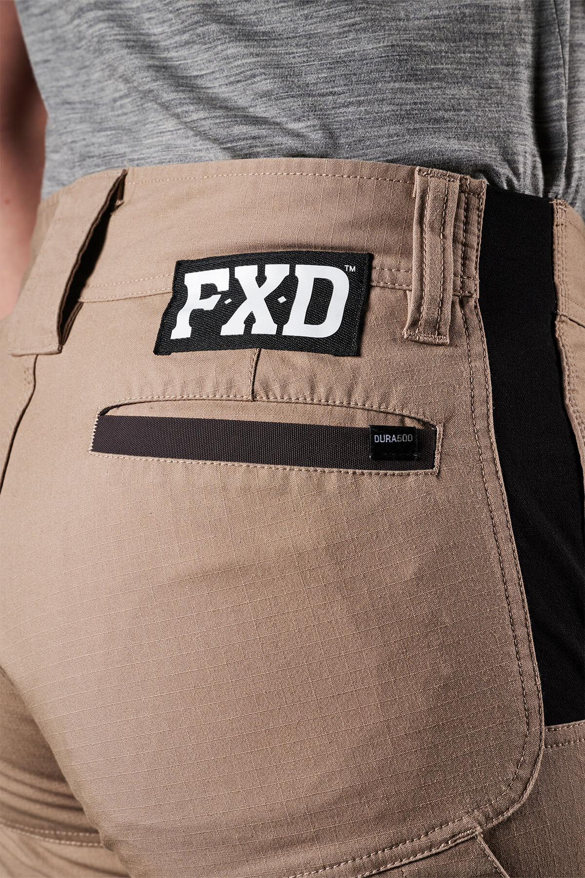 Close-up of a person wearing the FXD Womens WP-8W lightweight stretch ripstop pants in beige. The pants feature a black and white FXD logo patch on the back waistband, a black horizontal zippered pocket labeled "DURASCO" on the right side, and offer 50+ UPF protection. The person is also wearing a grey shirt.