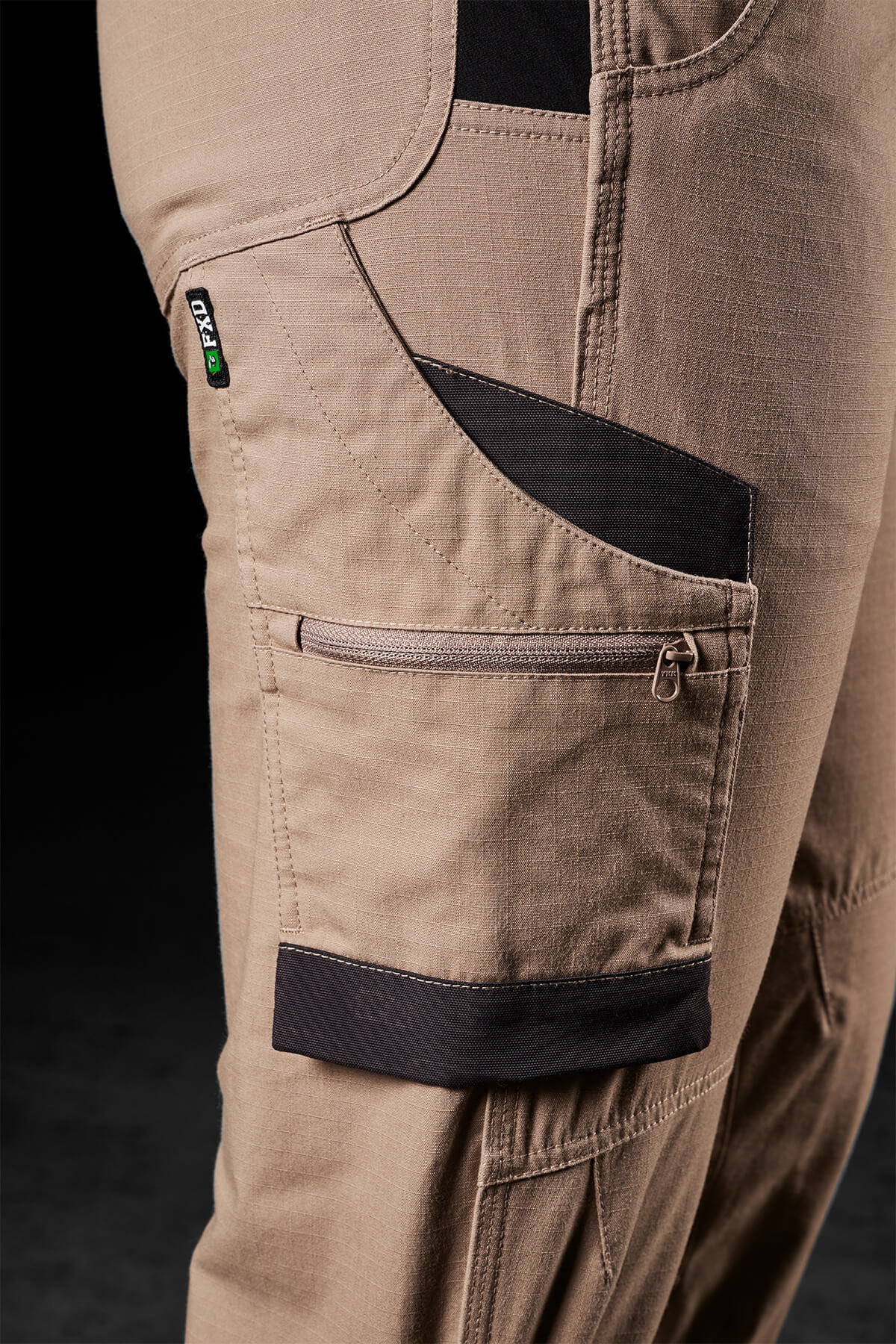Close-up view of a pair of FXD Womens WP-8W khaki work pants with a focus on the multi-functional side pocket featuring a zipper closure and black fabric accents. These lightweight stretch ripstop pants offer maximum range of movement, detailed with stitching, a small green logo tag, and sections for tools or accessories.