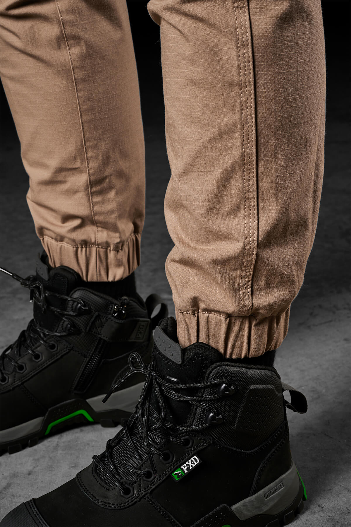 Close-up view of a person wearing FXD Womens WP-8W pants in beige, which offer 50+ UPF protection and are cuffed at the ankles. They are paired with black high-top work boots featuring green accents and the FXD logo on the side, set against a dark, textured background.