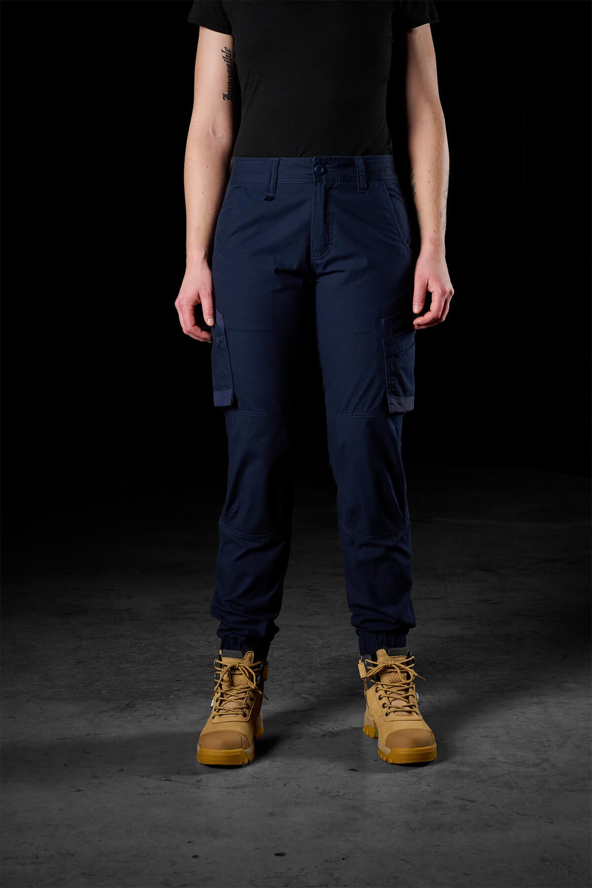 A person stands facing the camera, wearing a black top, FXD Womens WP-8W pants in blue, and tan-colored tactical boots. The dark background highlights the clothing's 50+ UPF protection and the sturdy footwear.
