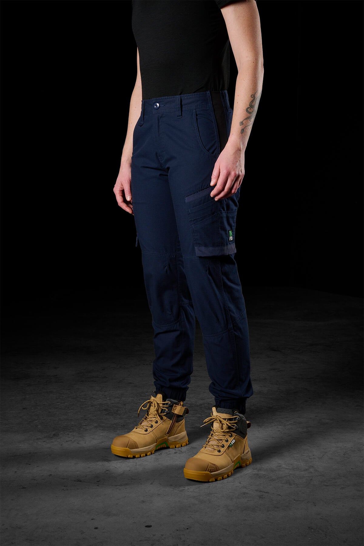 A person stands against a dark background, wearing the FXD Womens WP-8W stretch ripstop pant paired with a black shirt and tan work boots. Their right arm has a tattoo, and their hands are relaxed at their sides.