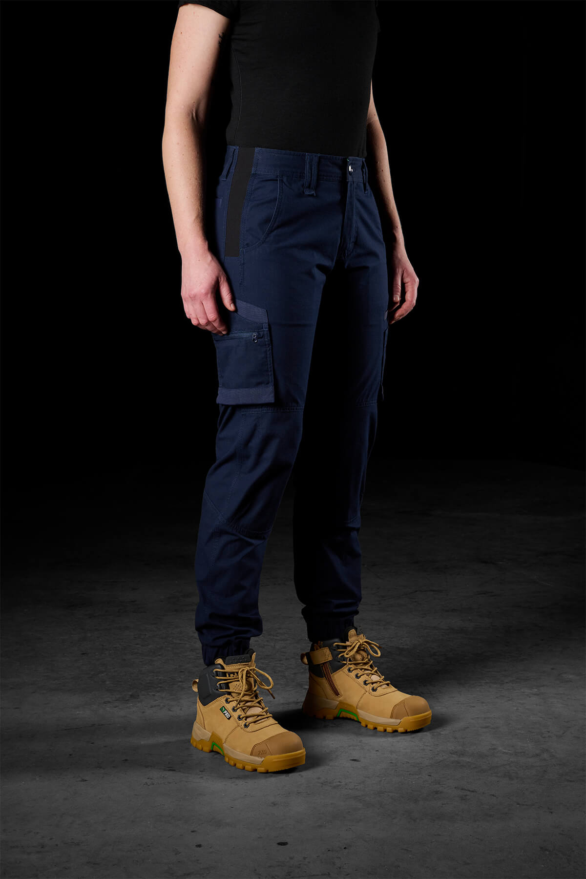 A person stands against a dark background, dressed in a black short-sleeve shirt, FXD Womens WP-8W stretch ripstop pants with 50+ UPF protection and a side pocket, and tan heavy-duty work boots. The photo is taken from shoulder to feet. The person’s arms are relaxed by their sides.