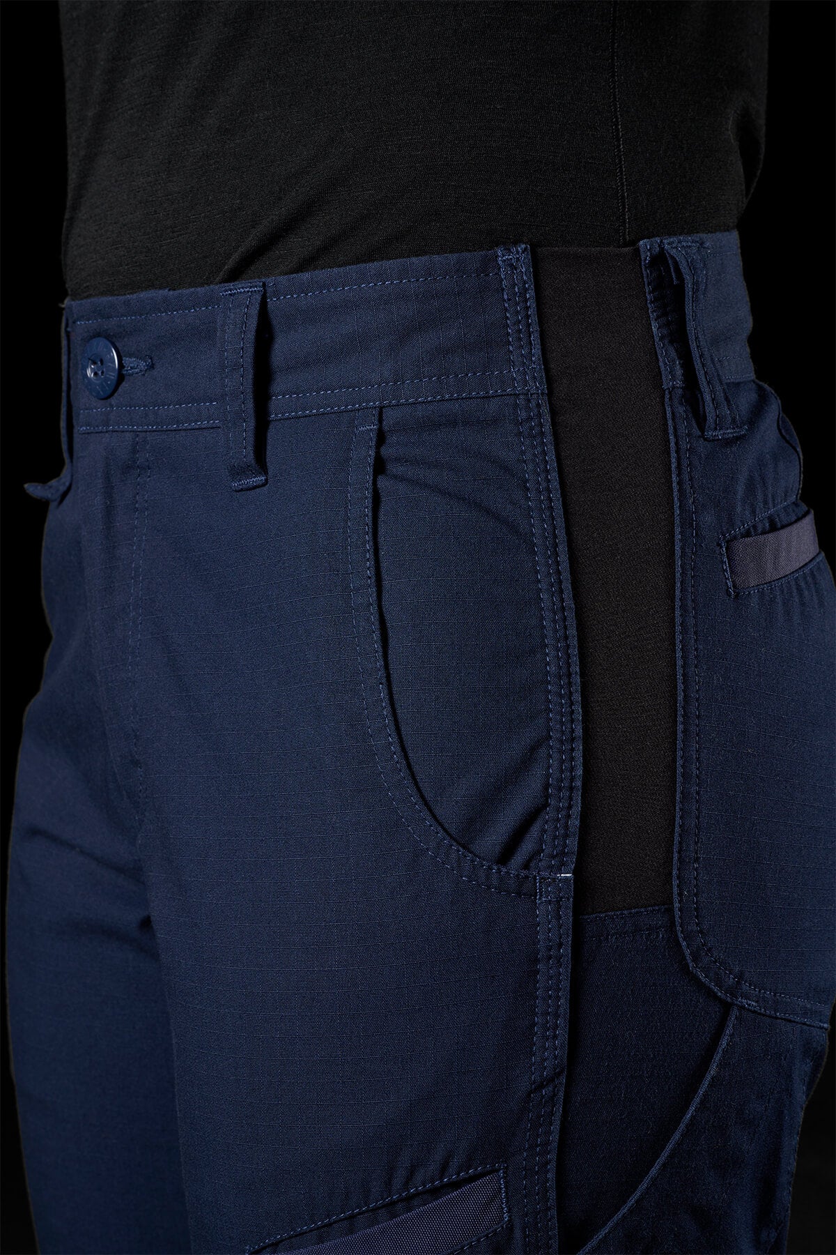 Close-up of a person wearing dark blue FXD Womens WP-8W stretch ripstop pants and a black shirt against a dark background. The FXD Womens WP-8W pants feature a diagonal side pocket, belt loops, button closure, intricate stitching details, and 50+ UPF protection. The person's side profile is partially visible from the waist down.