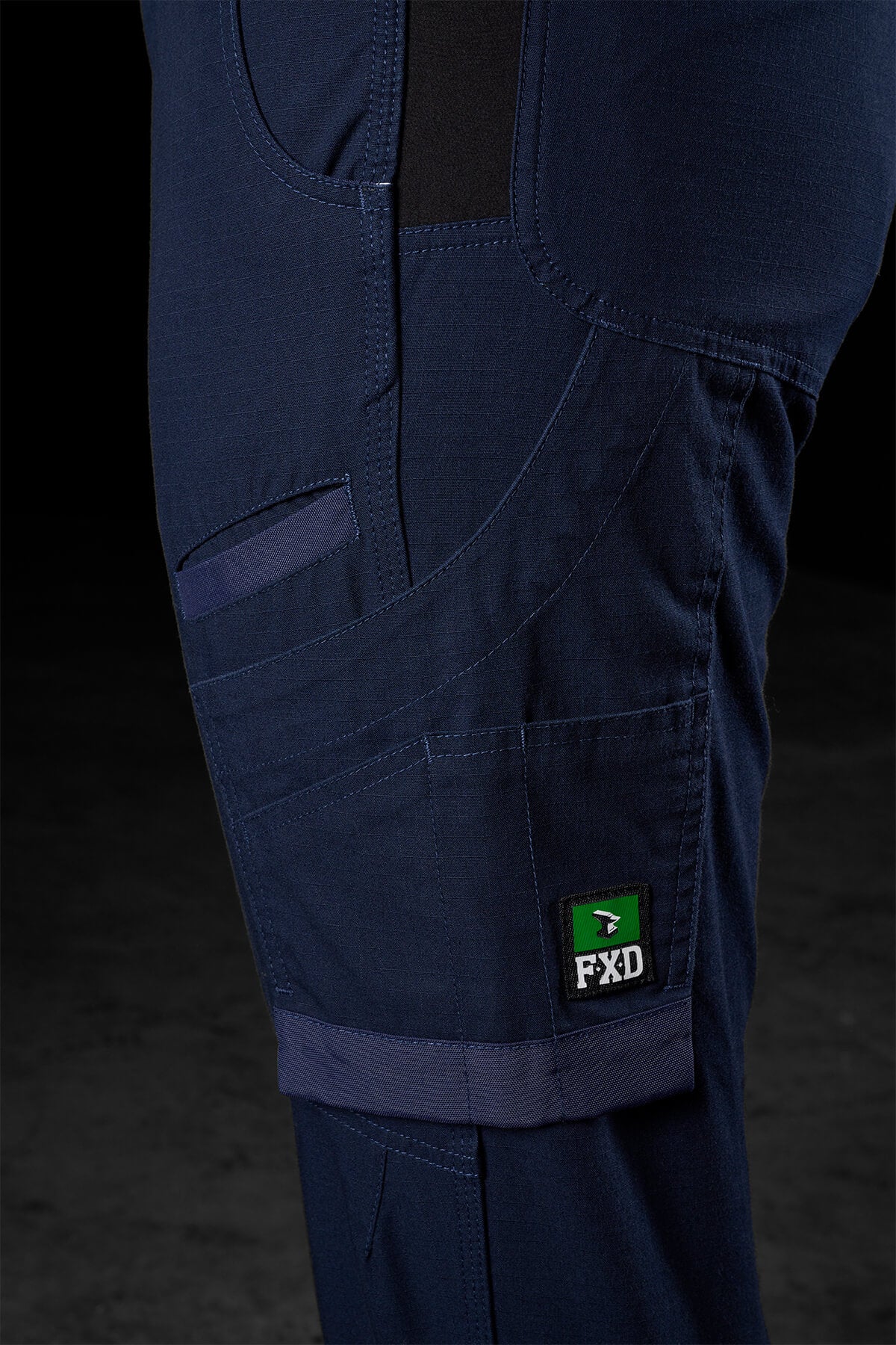 In the close-up of the pocket area of the dark blue FXD Womens WP-8W utility pants, you can see detailed stitching and multiple compartments. One pocket is accented with a green and white tag labeled "FXD". These stretch ripstop pants provide 50+ UPF protection. The background features a dark, out-of-focus surface.