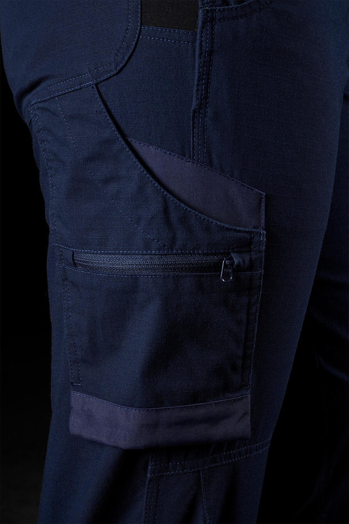 Close-up of a navy blue FXD Womens WP-8W stretch ripstop pant. The image highlights a side pocket with a zipper closure, featuring additional smaller compartments. The stitching and fabric texture are clearly visible, suggesting durability and functionality with 50+ UPF protection. The background is dark.