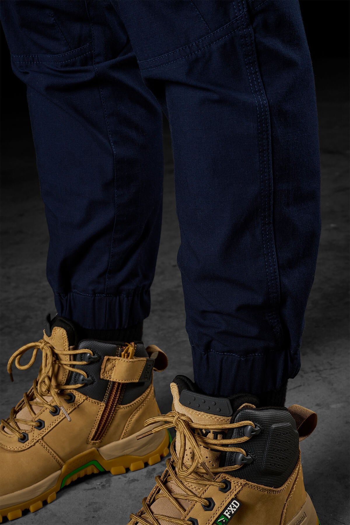 Close-up of a person wearing FXD Womens WP-8W stretch ripstop pants with elastic cuffs, paired with tan work boots featuring black and yellow accents. The boots have visible laces and side zippers. In the background, there is a dark, textured surface.