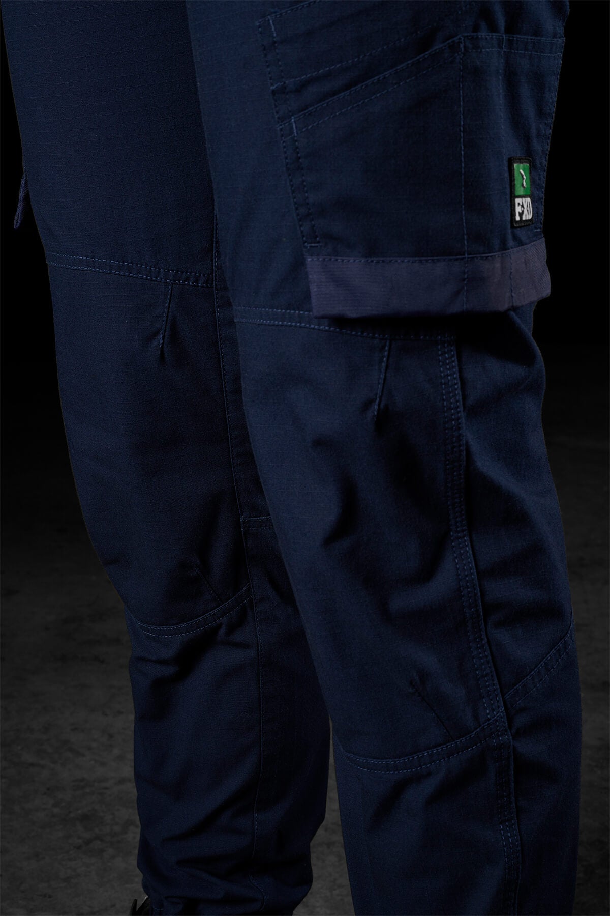 A close-up of the dark blue FXD Womens WP-8W cargo pants showcases their multiple pockets. These stretch ripstop pants feature a green logo tag on one thigh pocket and appear to be made of sturdy material. The dark background highlights the texture, details, and 50+ UPF protection of the pants.
