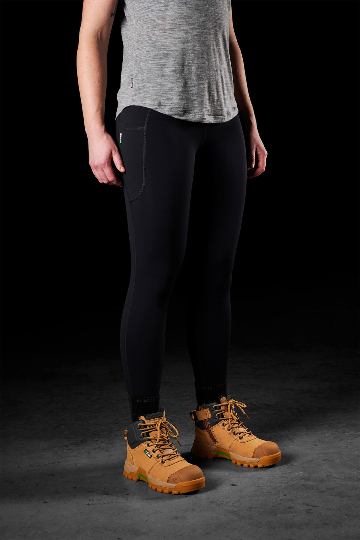 Person standing against a dark background wearing a gray sleeveless shirt, FXD Womens WP-9W Leggings with a high-rise waistband, and tan hiking boots. The individual's face is not shown. The outfit appears to be suited for outdoor activities.