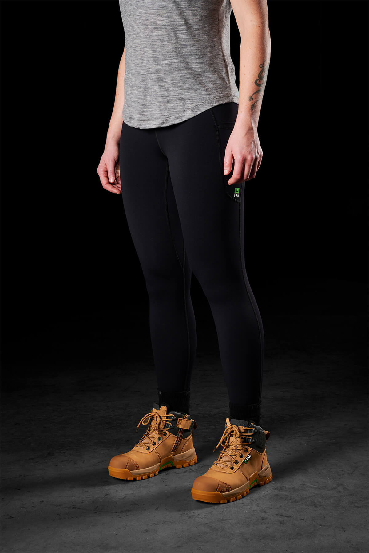 A person stands against a dark background, wearing a gray t-shirt, FXD Women's WP-9W leggings with a high-rise waistband, and tan work boots. Their arms are down by their sides, and their face is not visible. They have a tattoo on one forearm.