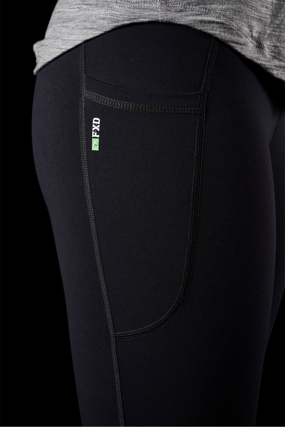 A detailed close-up displays a person donning FXD Womens WP-9W Leggings, highlighted by black athletic fabric, noticeable side stitching, and a discrete small pocket. An attached green and white tag reads "2FWD." These leggings boast a high-rise waistband and provide 50+ UPF protection against the backdrop of a black background.