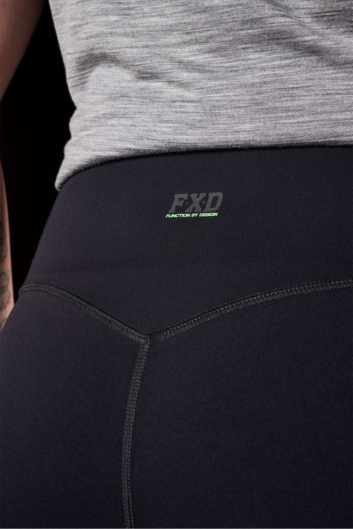 A close-up of an individual wearing FXD Womens WP-9W Leggings with a high-rise waistband, showcasing the visible seams and a small "FXD Function By Design" logo. The black athletic leggings provide 50+ UPF protection. Additionally, they are dressed in a light grey top and part of their tattooed arm is visible on the left side of the image.