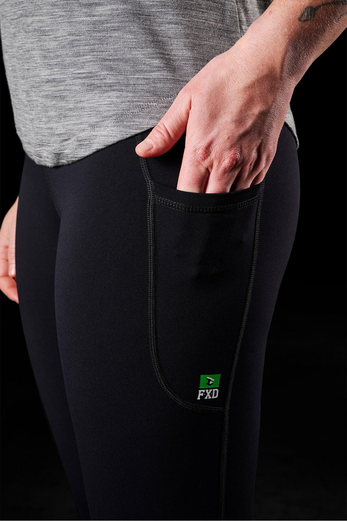 Close-up of a person wearing black FXD Womens WP-9W Leggings with 50+ UPF protection and a high-rise waistband. The leggings, which have a side pocket being held by the person's right hand, are paired with a gray top. The background is black and out of focus.