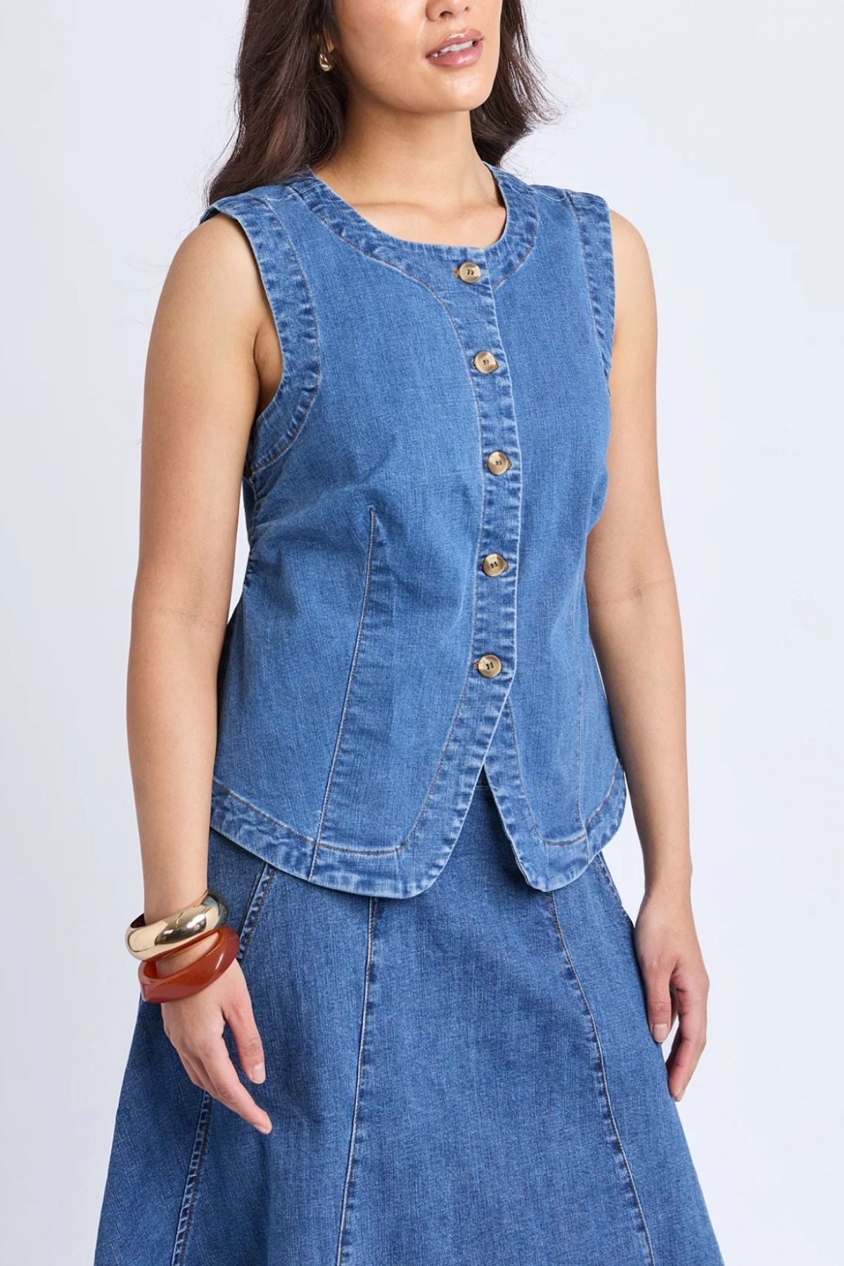 A woman models the Foxwood Abbott Vest in a modern silhouette, paired with a matching denim skirt and chunky bracelets on her left wrist, standing against a plain light background.